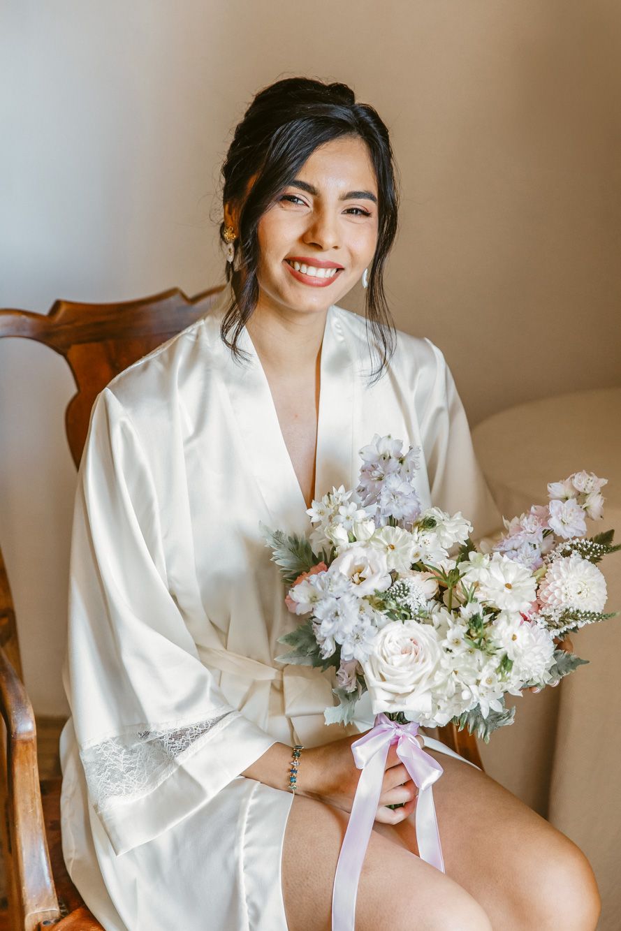 Smiling bride in white satin robe holding elegant bouquet of white and pink flowers while seated on wooden chair.