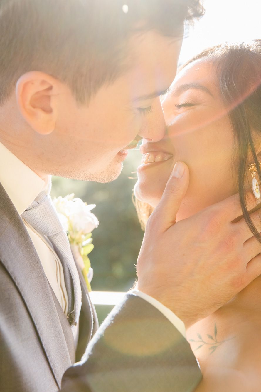 A groom in a gray suit tenderly holds his smiling bride's face as they share a romantic kiss in golden sunlight.