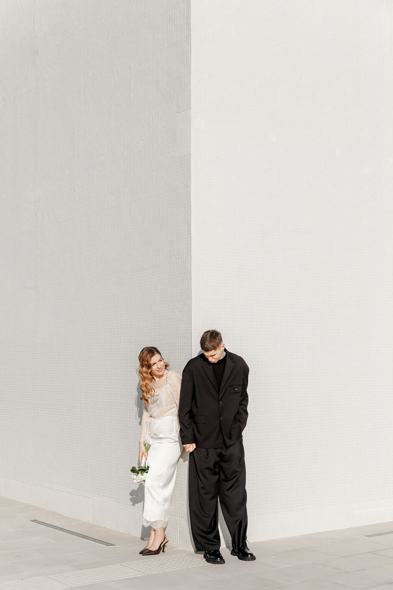 Bride in white outfit and groom in black suit leaning against a white wall, holding a small bouquet.