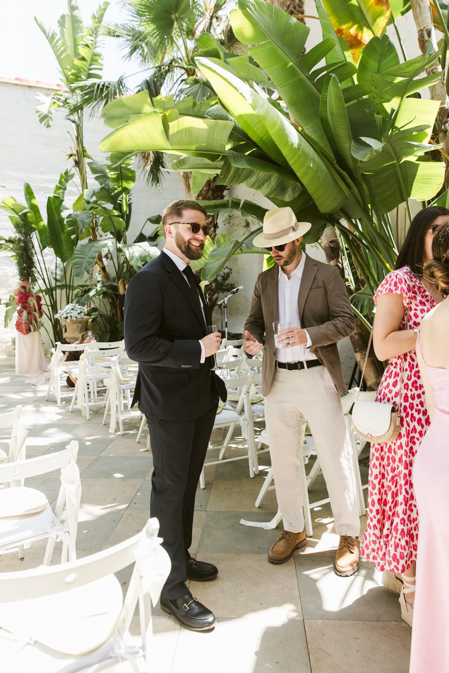 Two well-dressed men socializing at an outdoor wedding venue surrounded by tropical plants and white chairs.