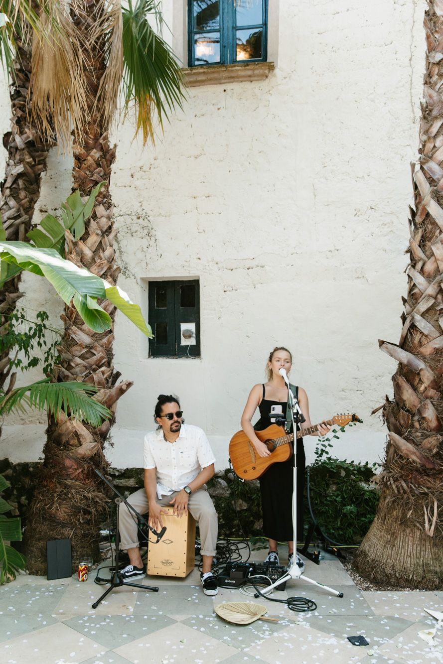 Acoustic duo performs outdoors with guitar and cajon between palm trees against a white stucco wall.