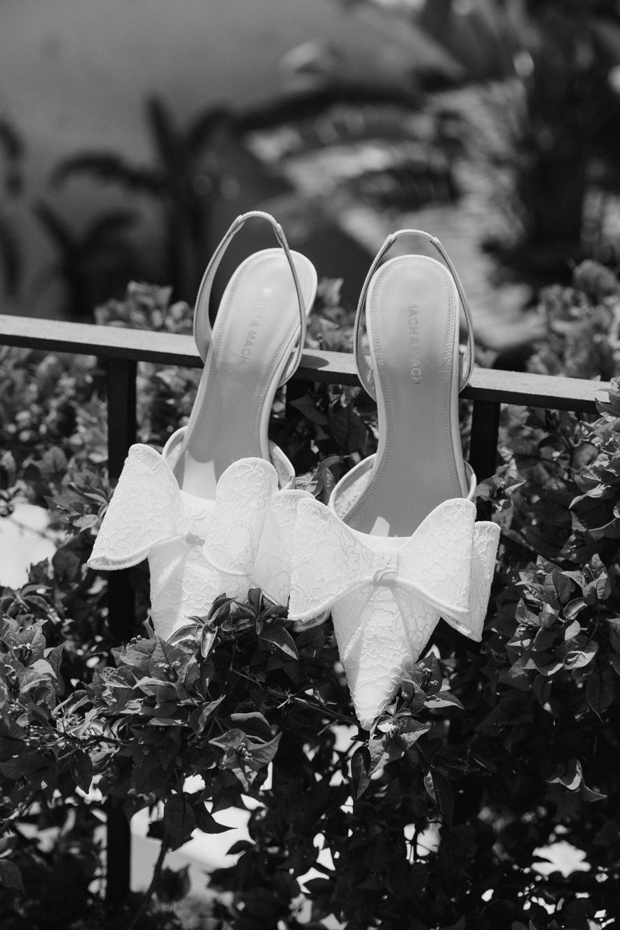 Black and white photo of elegant bridal heels with lace bow detail resting on garden foliage.