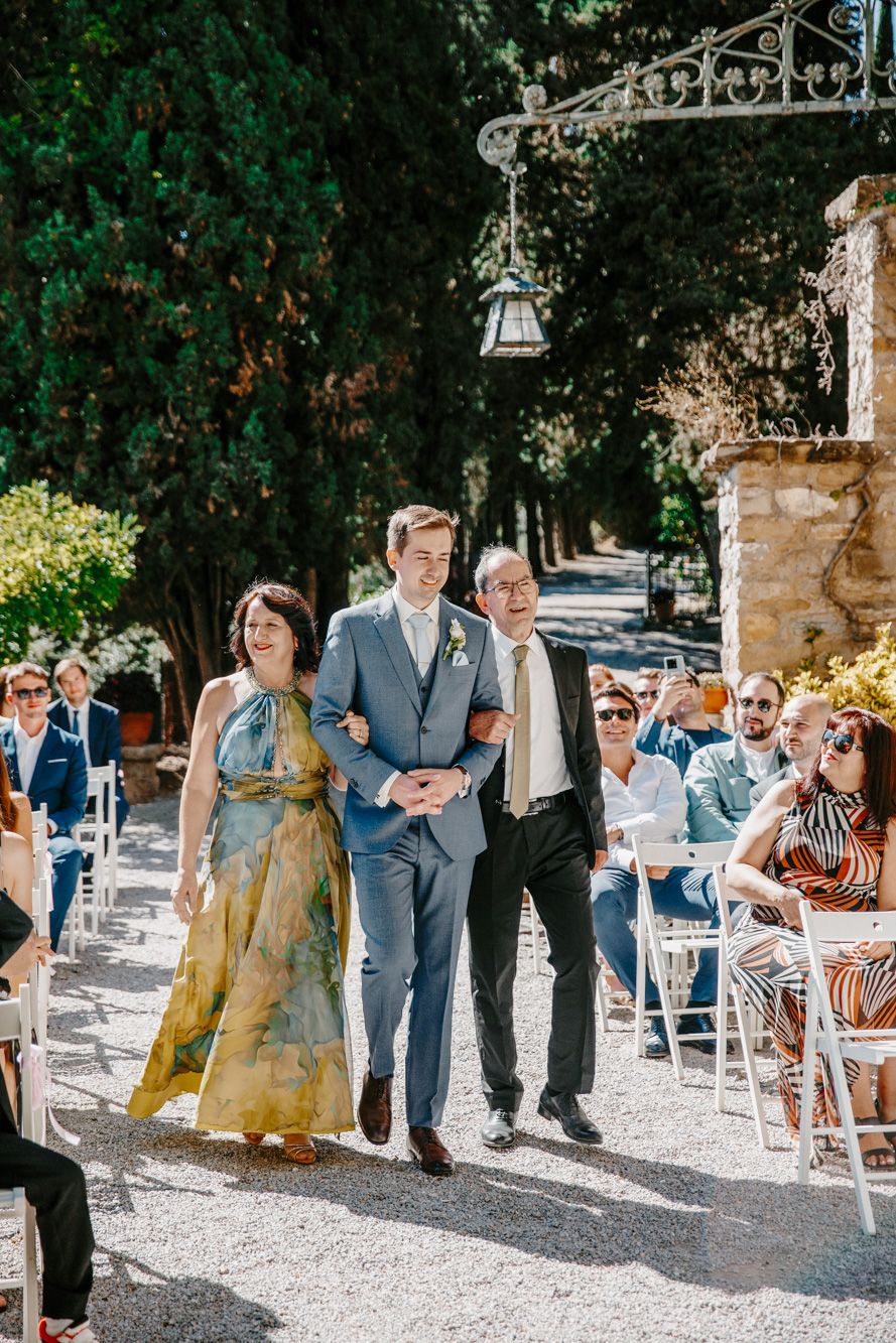 Groom walks down outdoor wedding aisle escorted by parents, guests seated on white chairs, sunny garden venue.