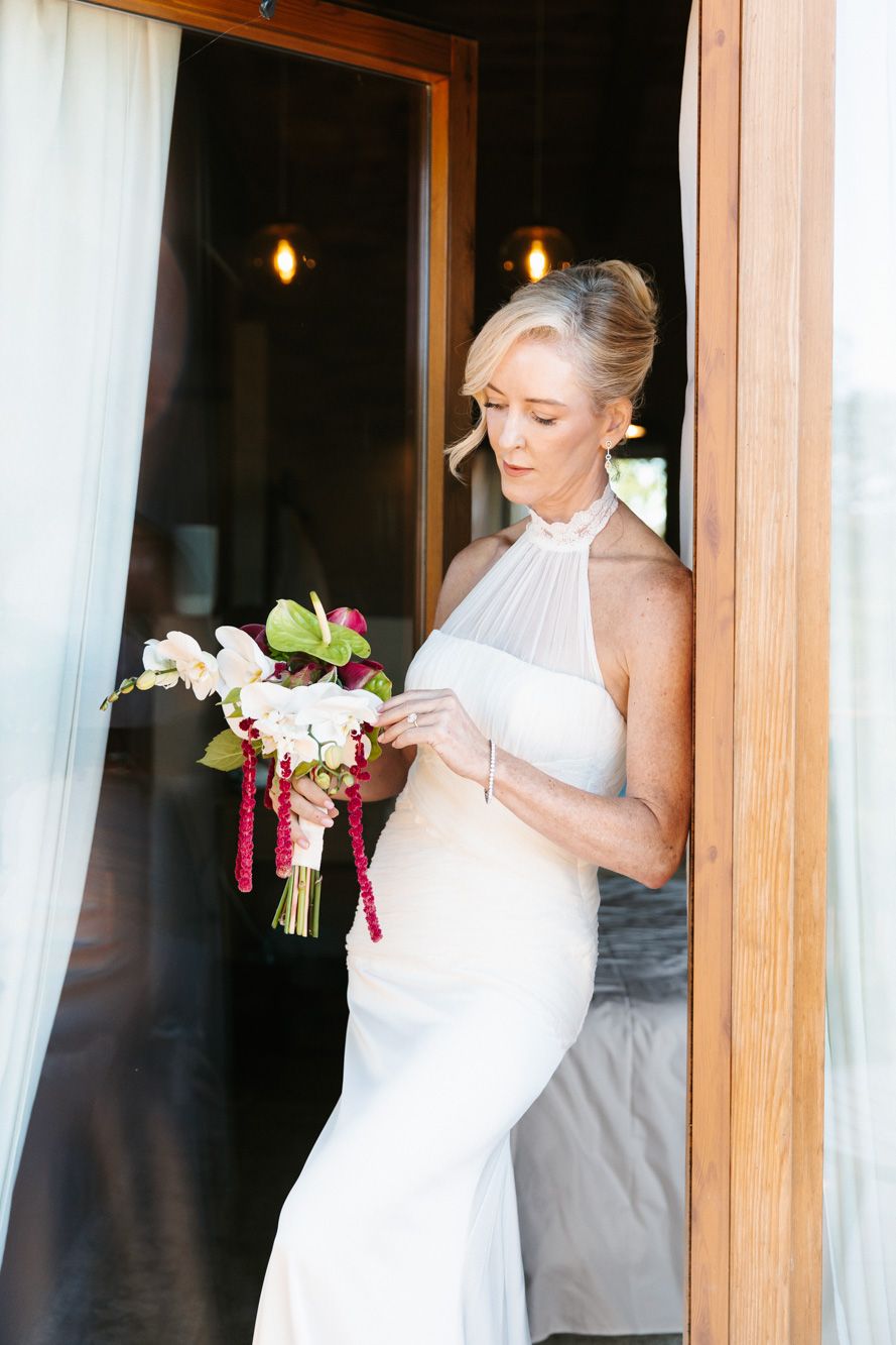 Bride in a white halter-neck wedding gown holds a bouquet of white flowers and red beaded accents while leaning against a wooden doorway.