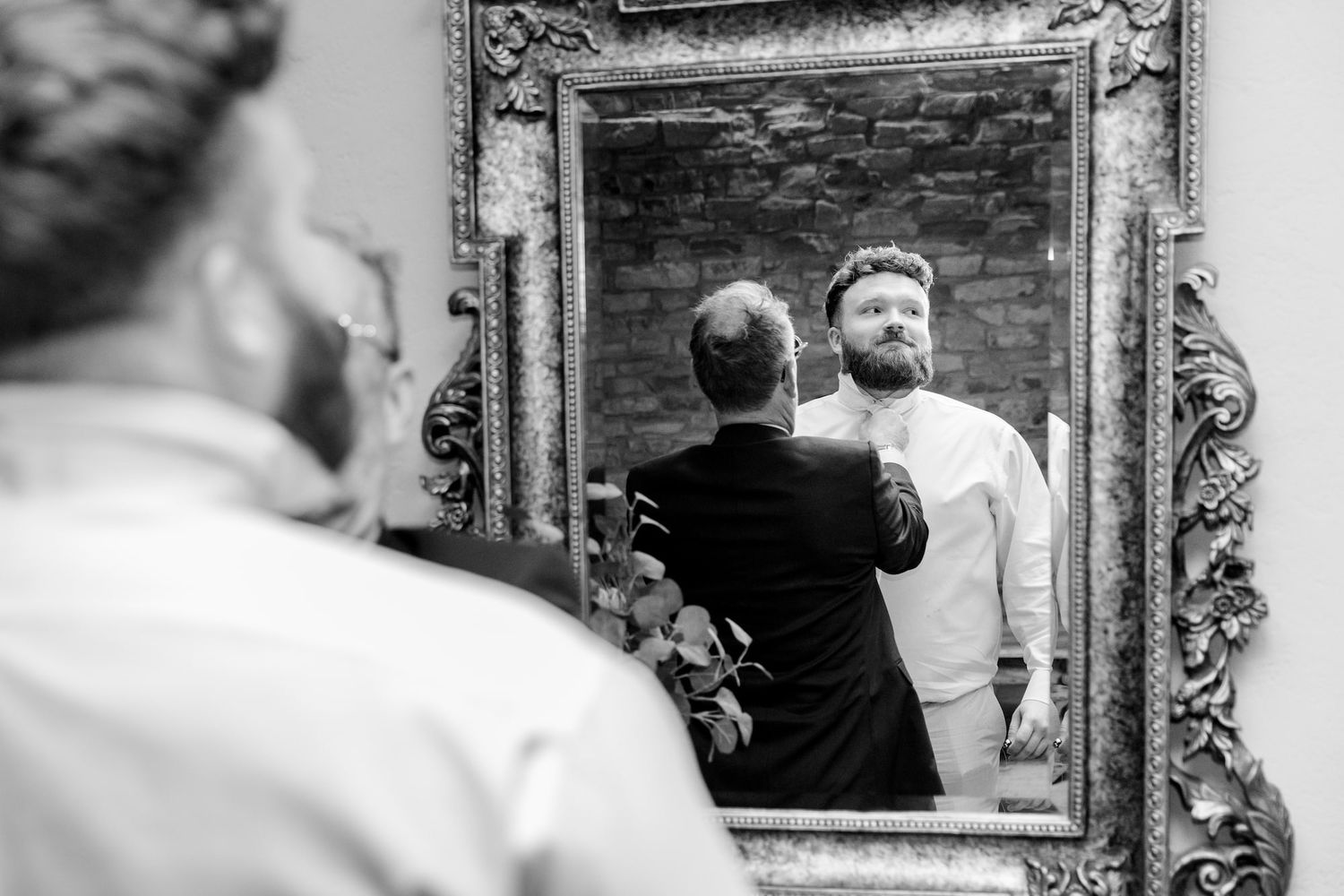 Black and white photo of a man being helped with his shirt in front of an ornate mirror.