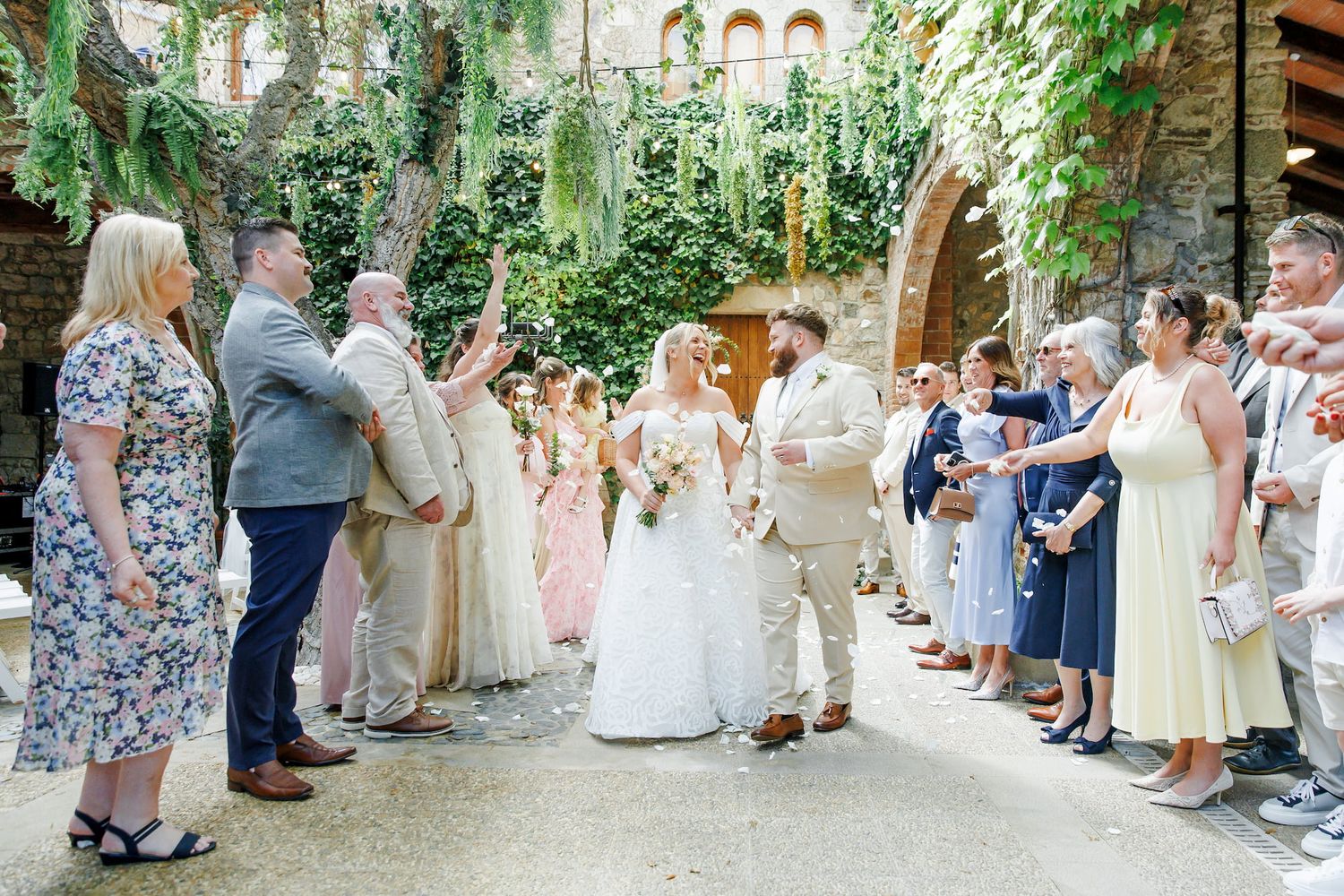 Bride and groom walk through cheering wedding guests at an outdoor ceremony venue with stone arches and lush greenery.