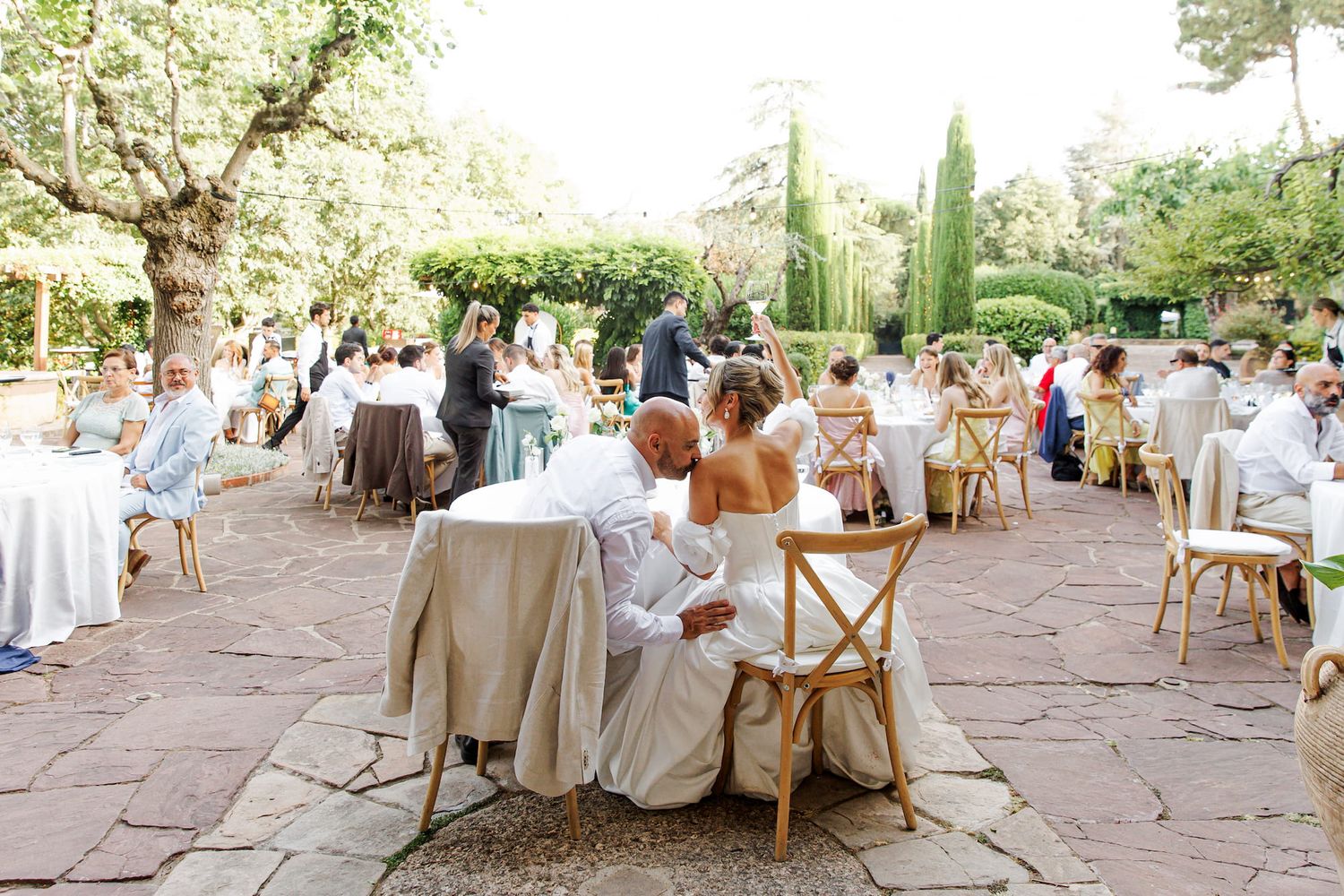 Wedding reception guests seated at white-clothed tables on stone patio surrounded by lush greenery and tall cypress trees.
