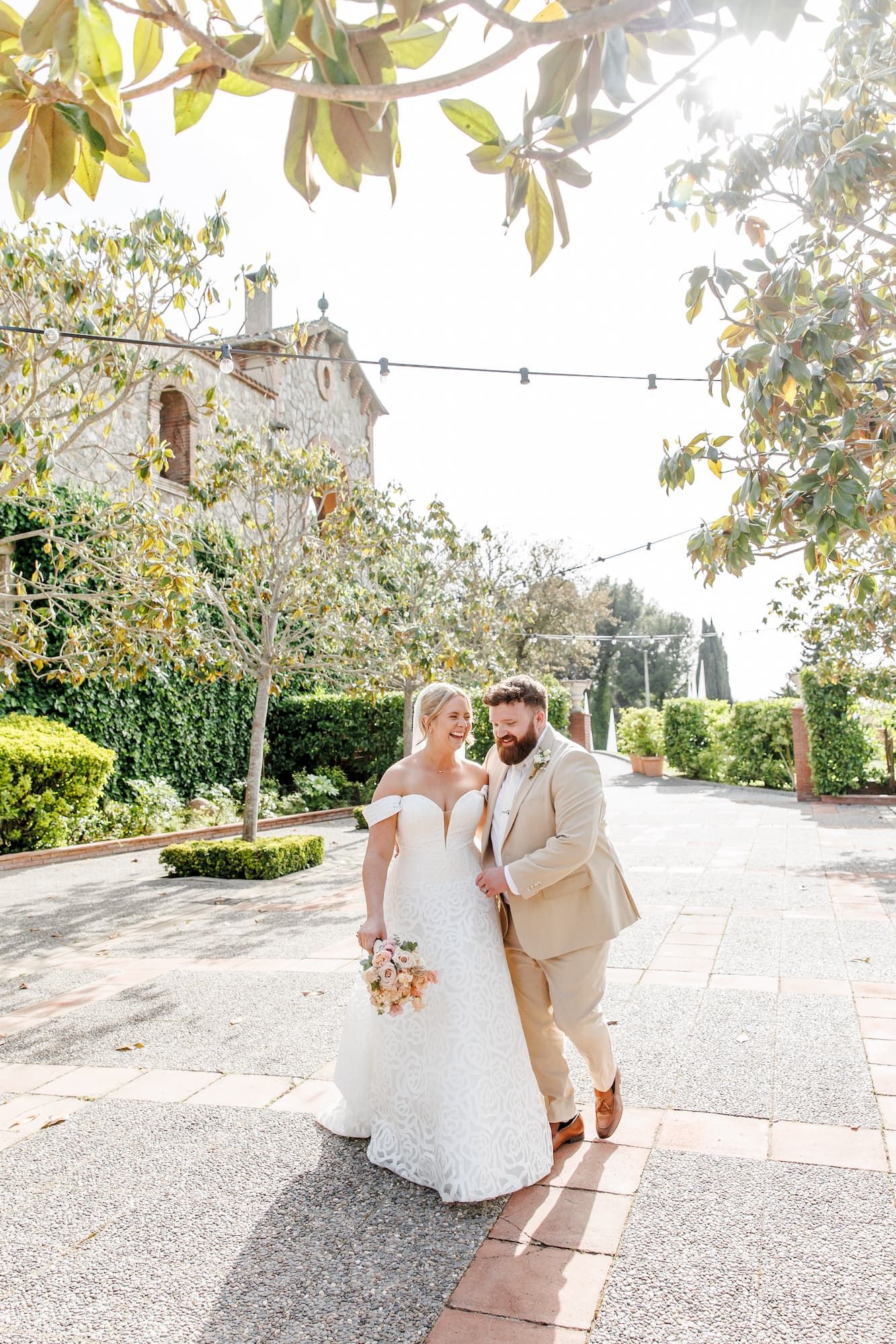 Bride and groom walking together outdoors at a sunlit garden of Mas de Sant Leli, she holds a bouquet in a white lace gown.