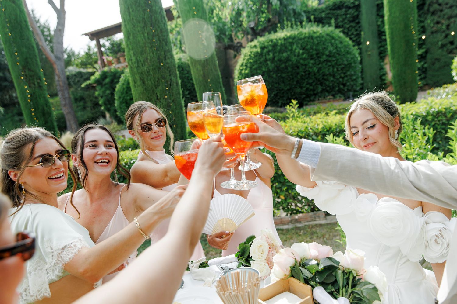 Women in white dresses toasting with Aperol Spritz cocktails outdoors in a lush green garden setting.