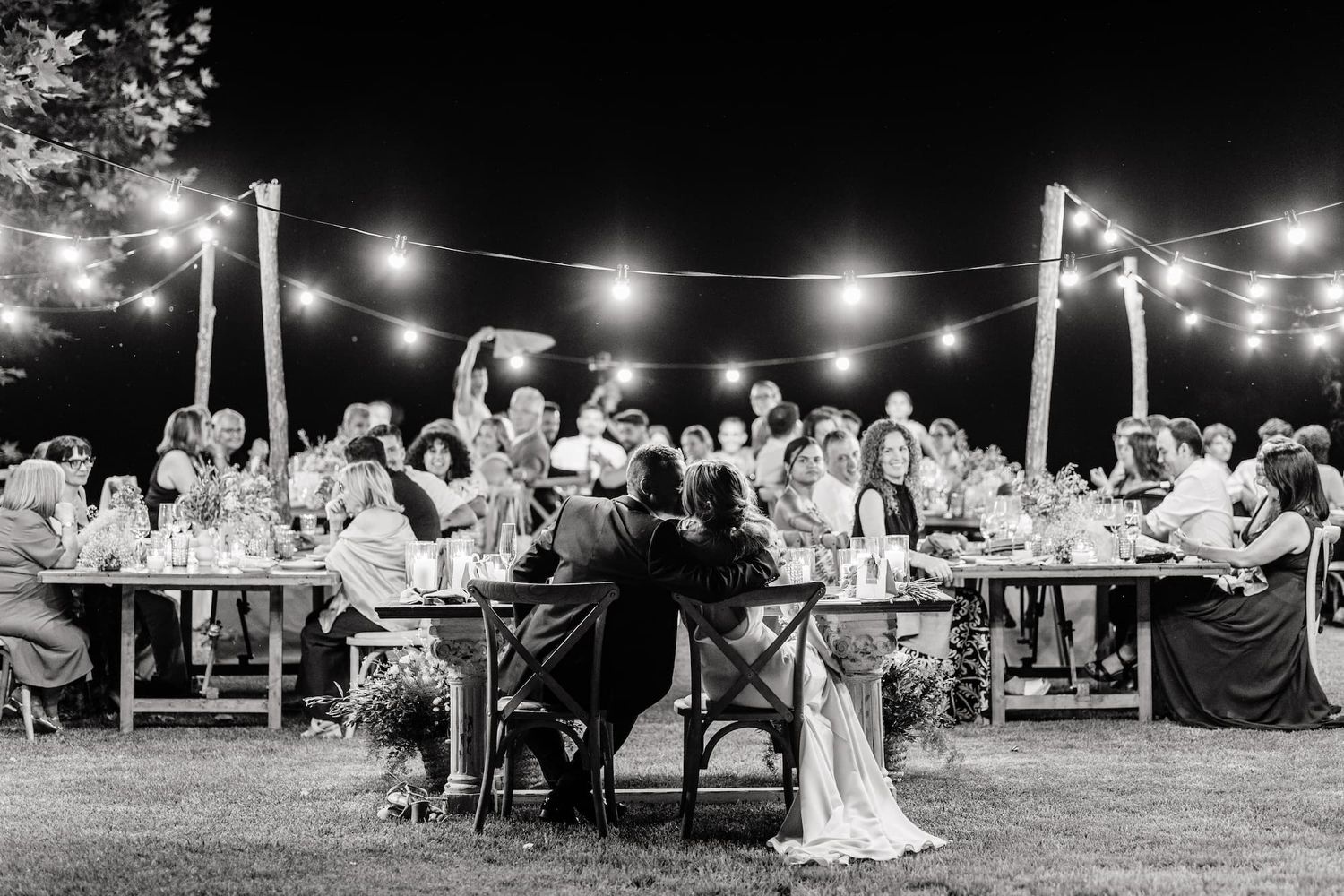 Black and white outdoor wedding reception under string lights with guests seated at long tables on a lawn at night.