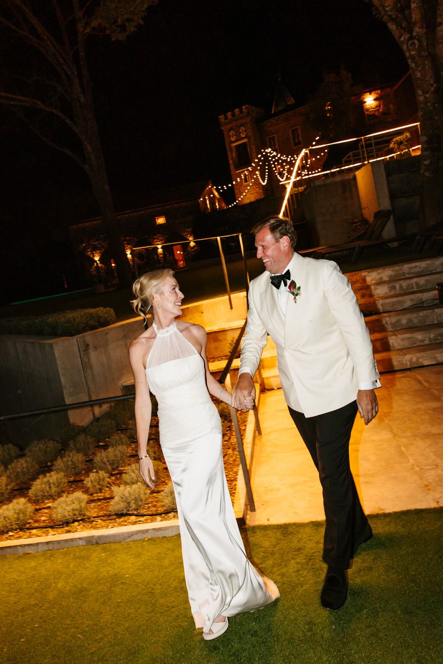 A bride in a white halter gown holds hands with a groom in a white dinner jacket, walking outdoors at night near a lit castle-style venue.