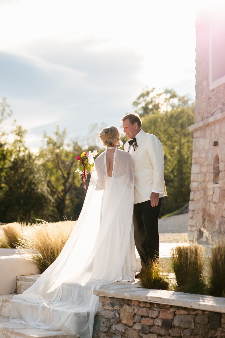 Bride in a flowing white gown with a long cape veil and groom in a white dinner jacket stand together on stone steps at sunset.