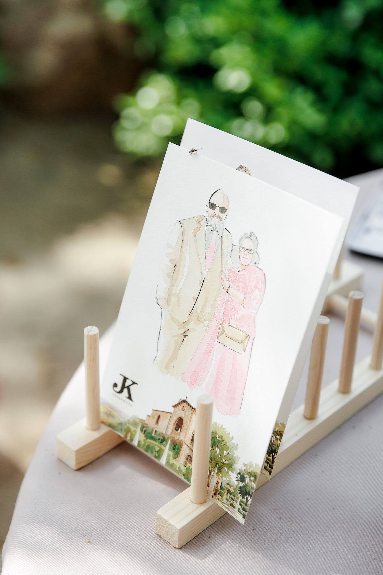 Watercolor illustration of a couple displayed on a wooden easel at an outdoor wedding reception.