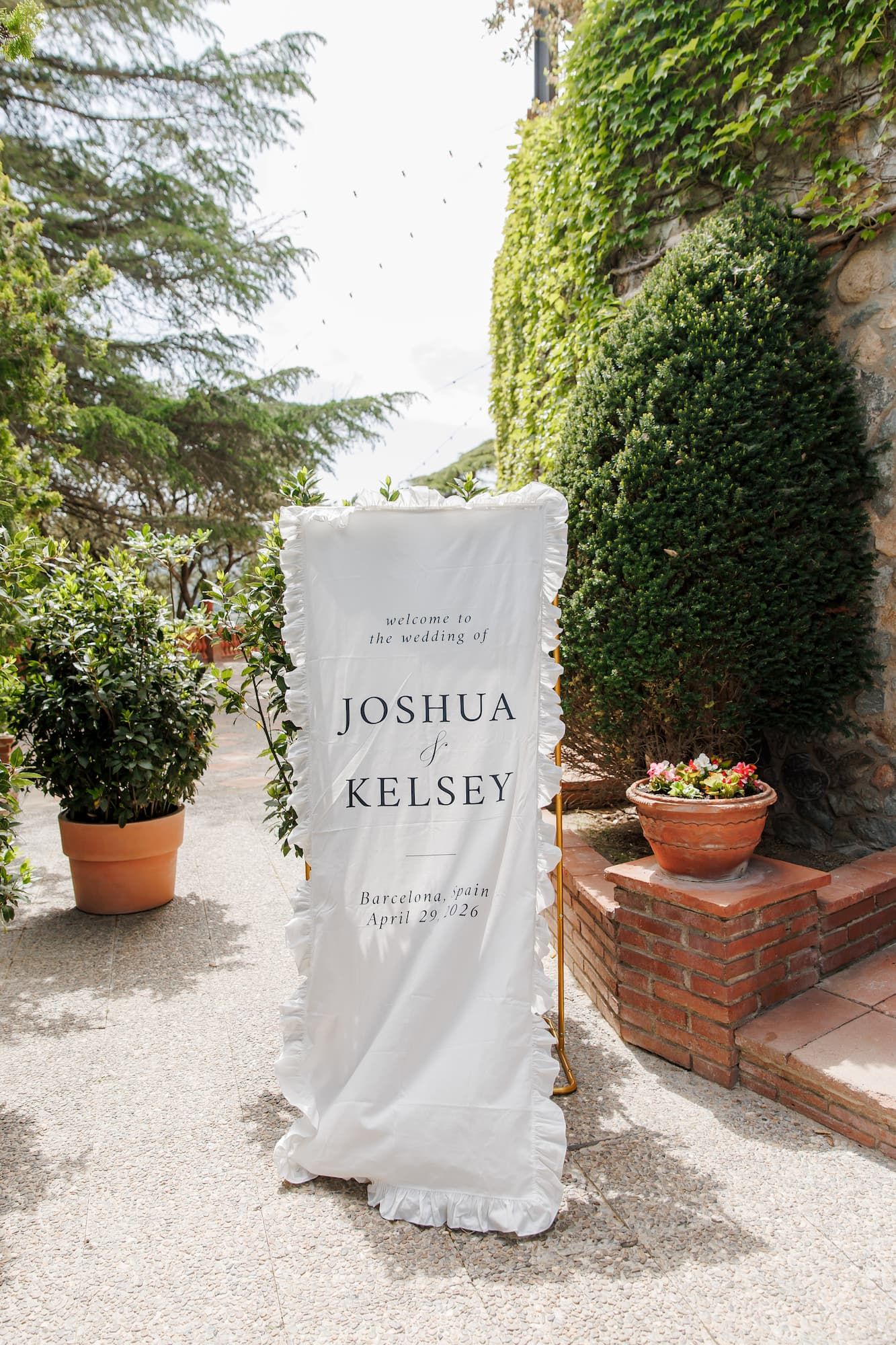 Elegant outdoor wedding welcome banner for Joshua and Kelsey in Barcelona, displayed on white fabric amid lush greenery.