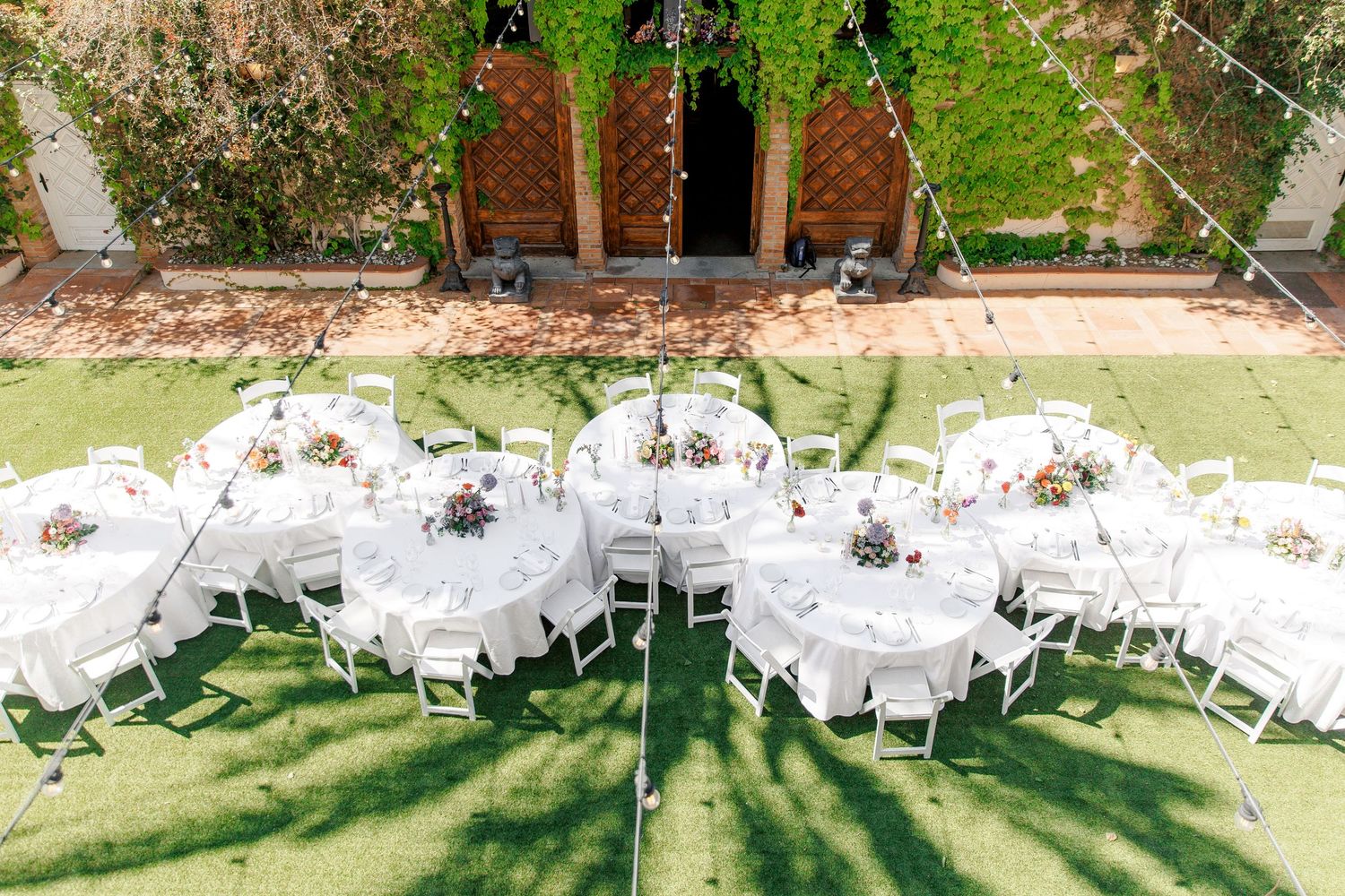 Outdoor wedding reception with round tables, white chairs, and floral centerpieces on a green lawn with ivy-covered walls.