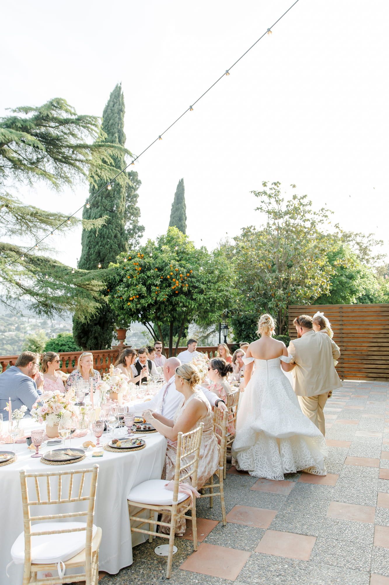 Elegant outdoor wedding reception with guests seated at round tables, bride in white gown, surrounded by lush greenery.