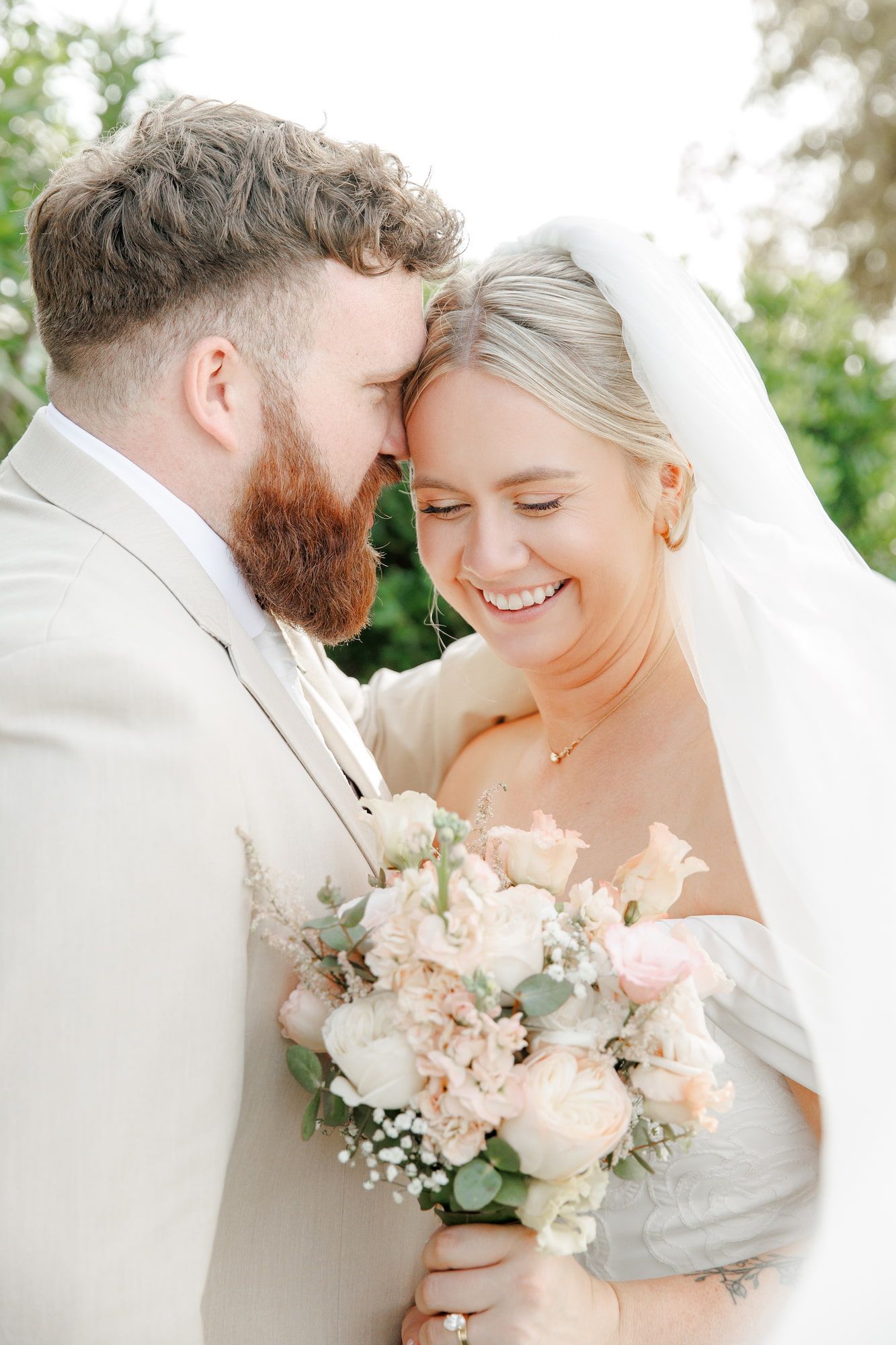 Smiling bride and bearded groom in beige suit share a tender moment, bride holding a blush bouquet.