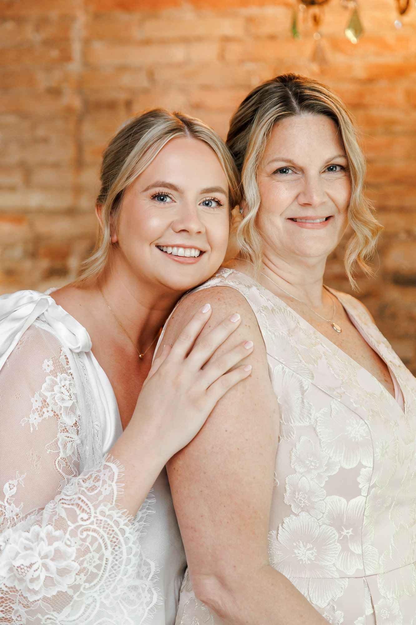 Two women in white lace wedding dresses smile together against a rustic brick wall backdrop.