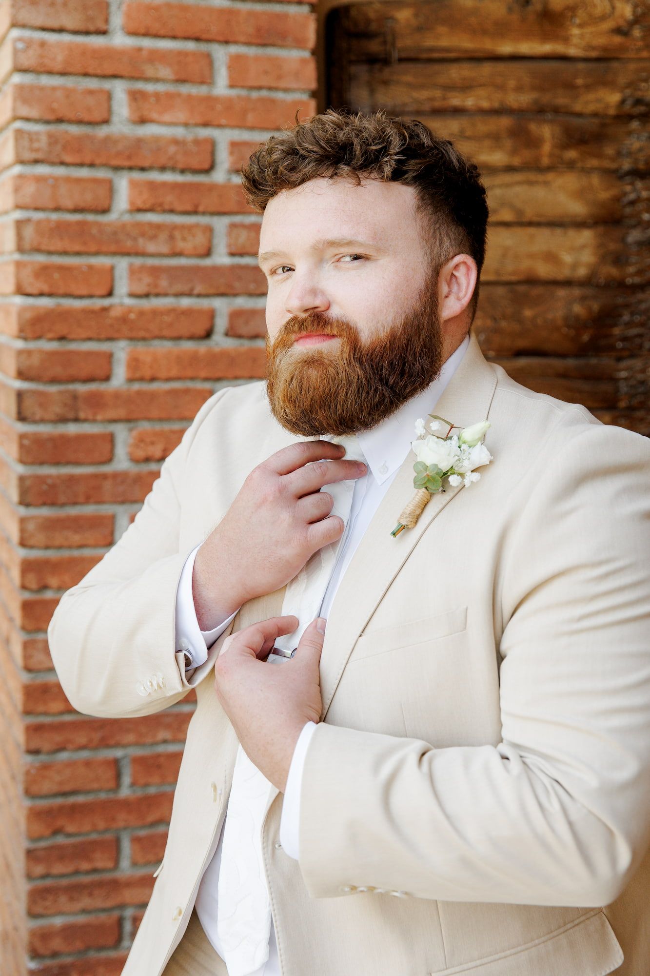 Handsome groom in cream suit adjusting tie with floral boutonniere against rustic brick wall.