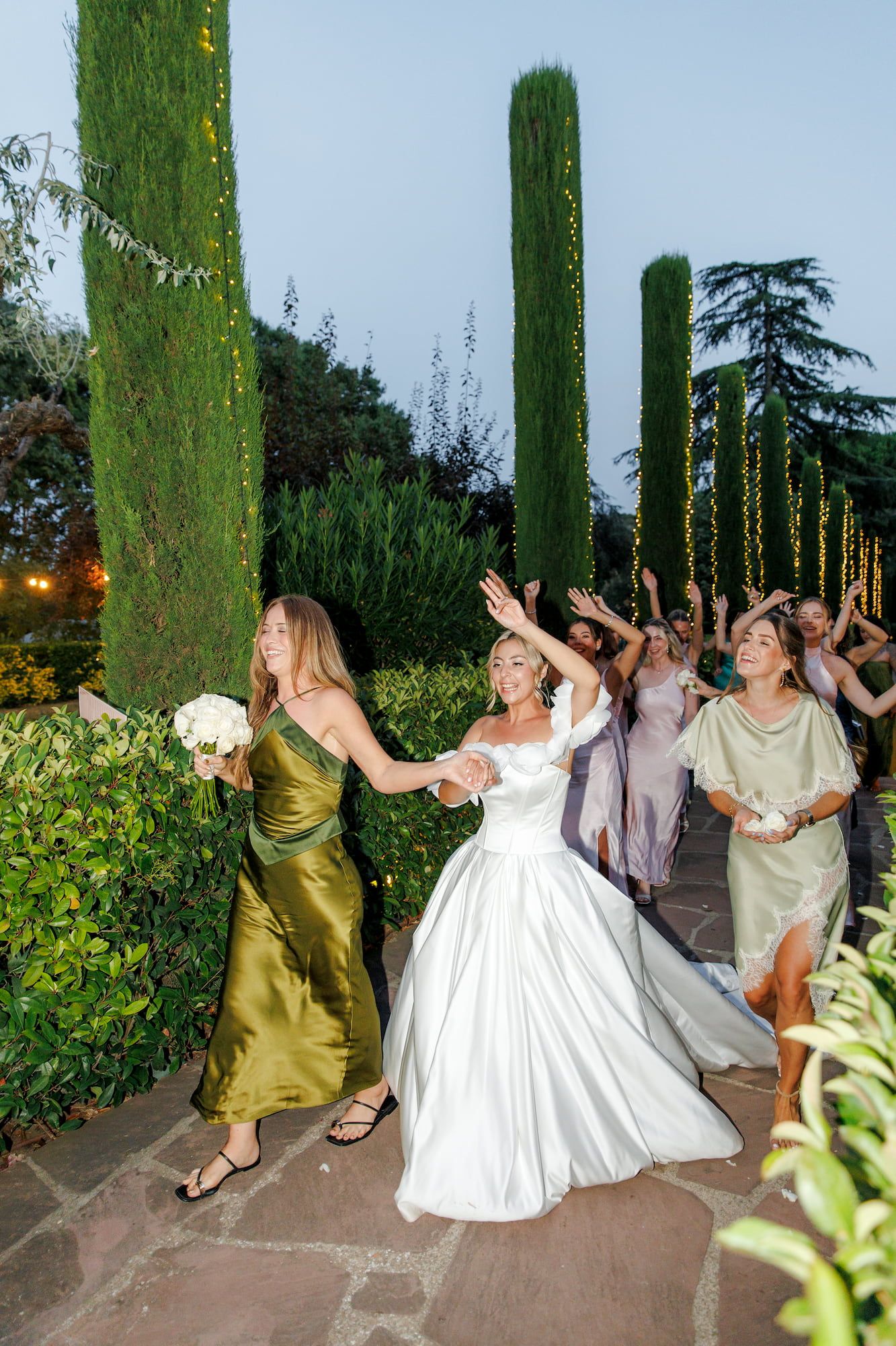 Bride in white gown walks with bridesmaids in colorful dresses through a garden with tall cypress trees.