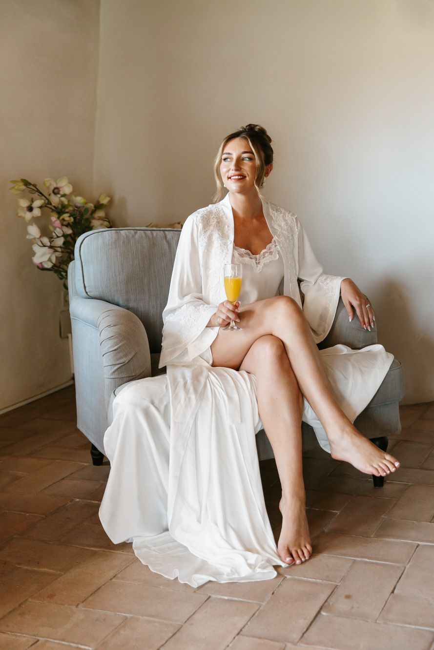 Smiling bride in white silk robe holding mimosa champagne flute, seated in gray armchair on wedding morning.