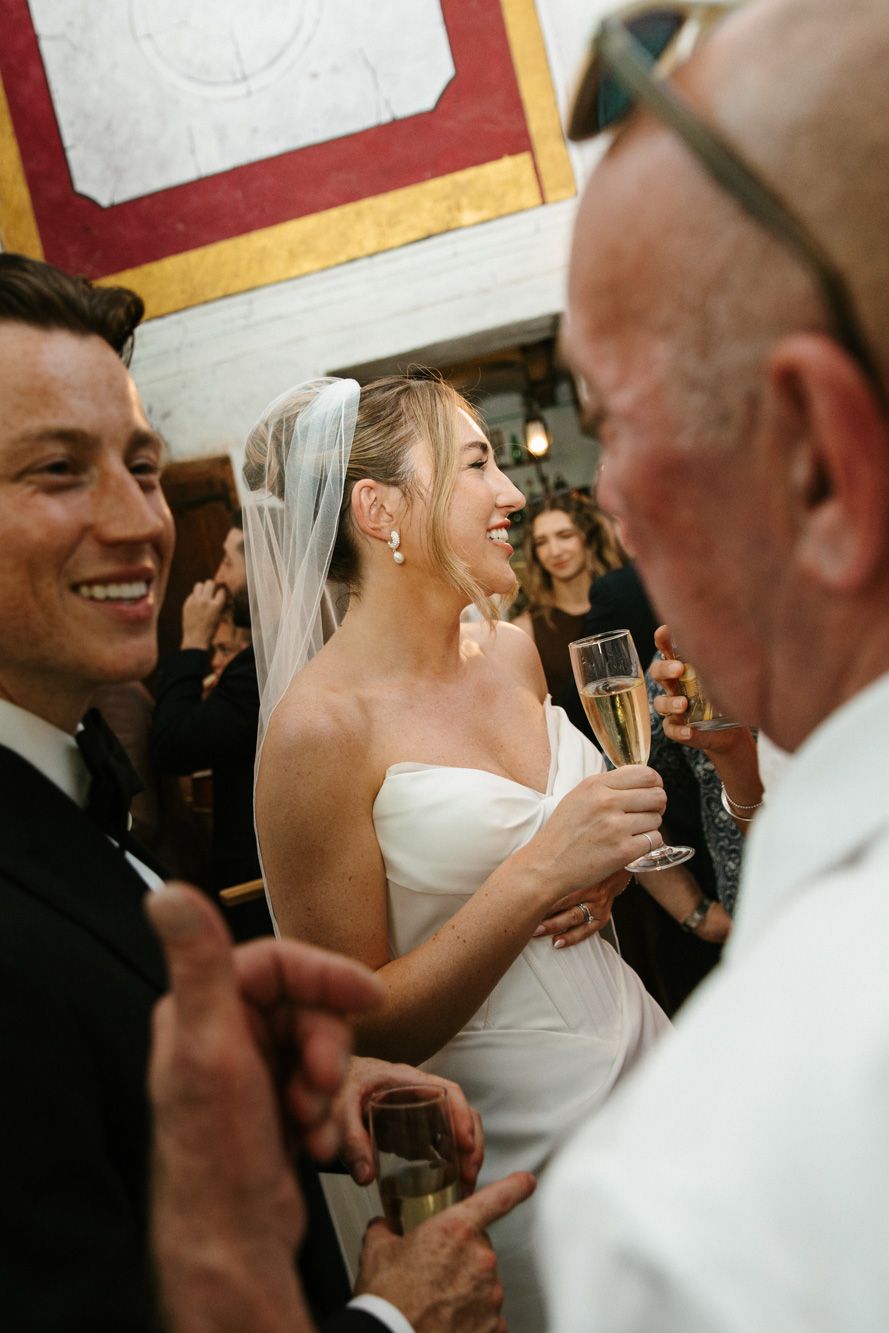 Bride in white gown and veil holds champagne flute, laughing with groom in black tuxedo at wedding reception.