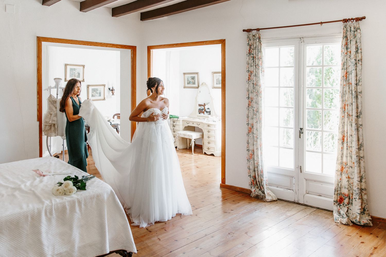 Bride in white gown with bridesmaid holding veil in bright rustic room with floral curtains and wooden floors.