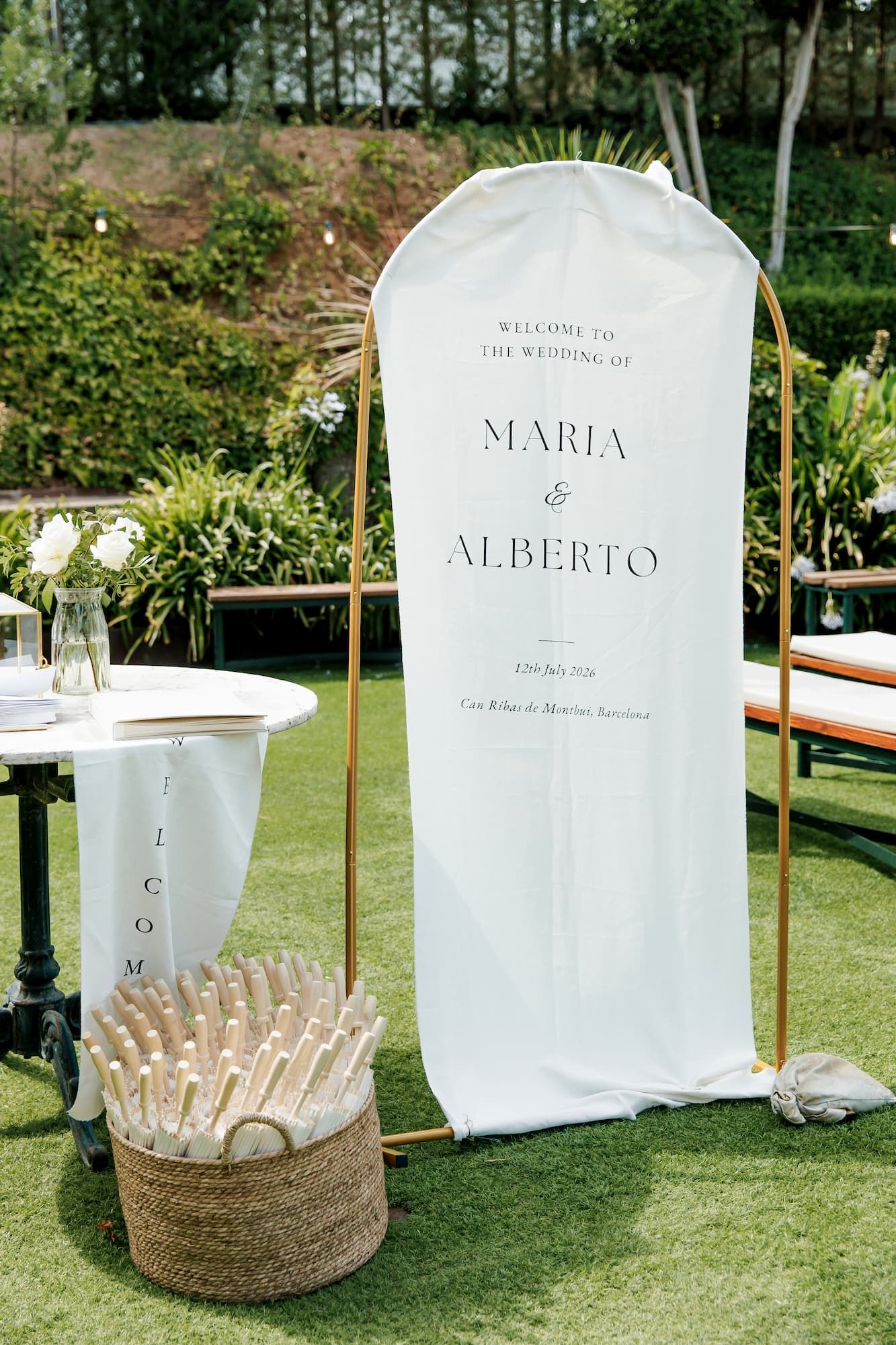 Elegant outdoor wedding welcome sign for Maria and Alberto on a gold arch stand with a wicker basket on green lawn.