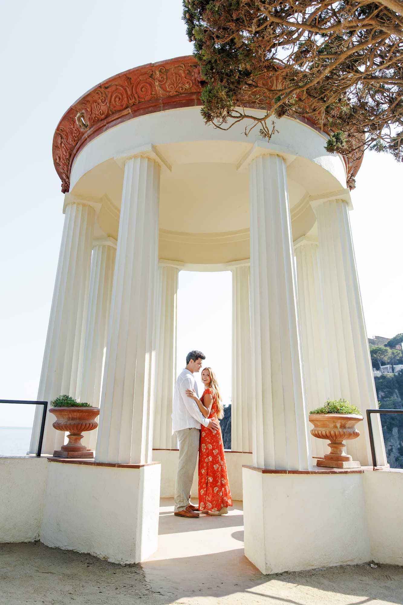 Couple embracing in a white classical rotunda with columns, woman in red floral dress, Mediterranean setting.