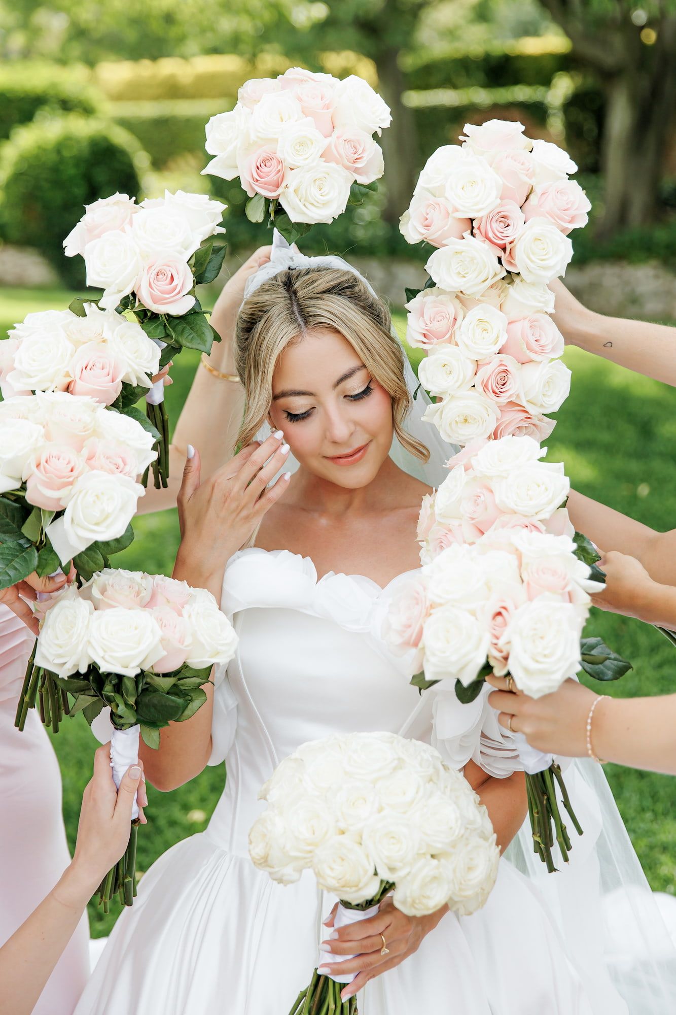 Bride in white gown holds bouquet of white roses, surrounded by bridesmaids with pink and white rose bouquets outdoors.