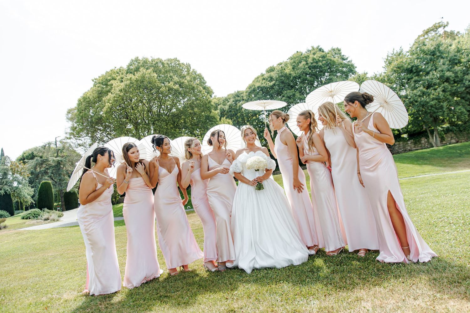 Bride in white gown with bridesmaids in blush pink dresses holding parasols on green lawn at outdoor wedding.