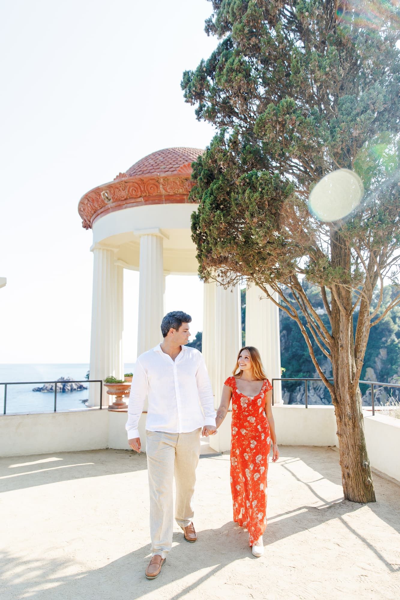 Couple holding hands walking by a Mediterranean gazebo, woman in red floral dress, man in white shirt, ocean view backdrop.