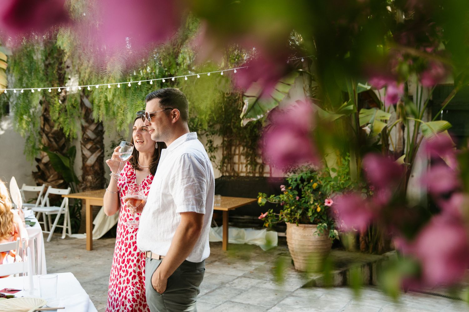 Couple embracing at an outdoor garden party with string lights, lush greenery, and pink flowers in the foreground.