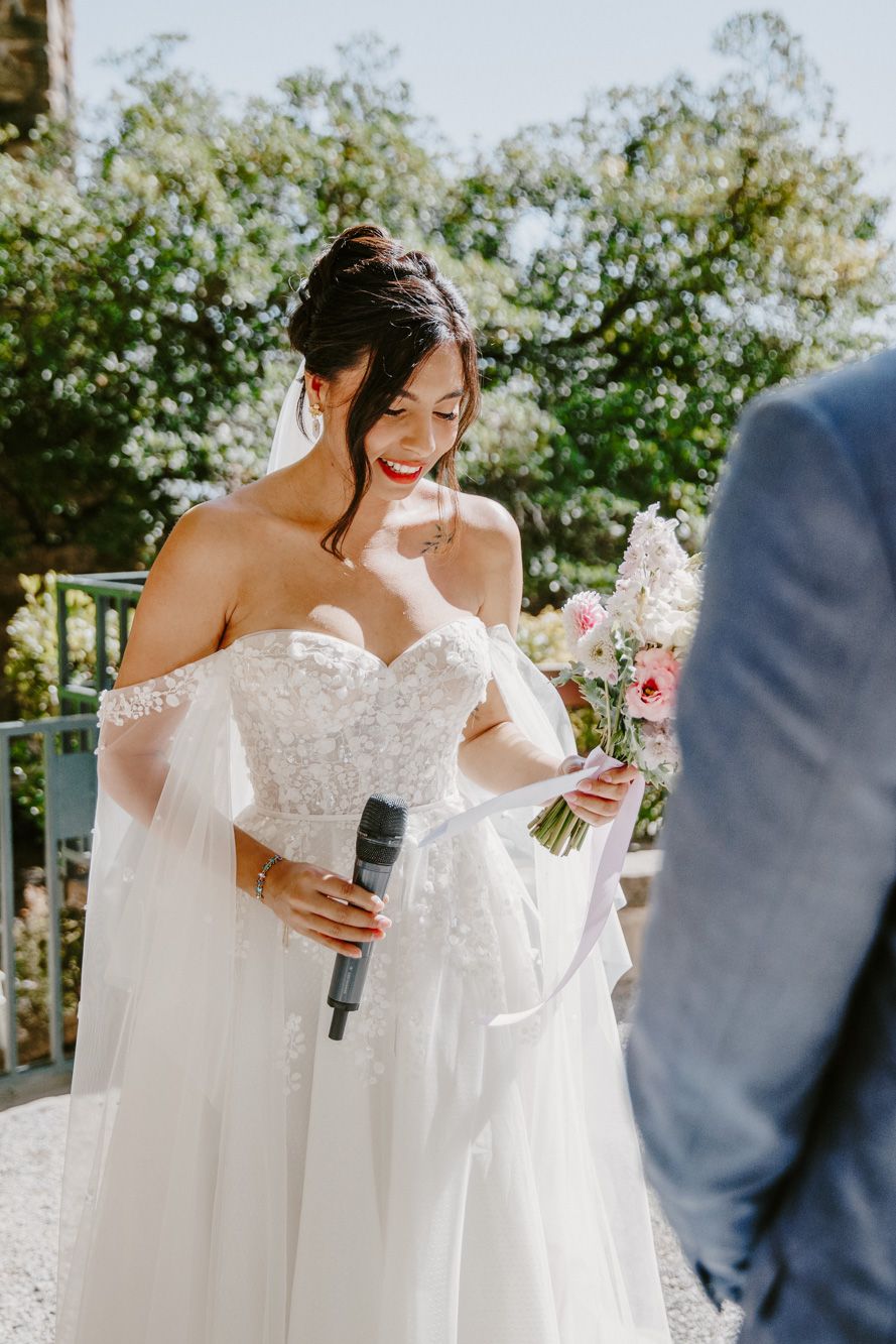 Smiling bride in off-shoulder lace gown holds microphone and pink bouquet during outdoor wedding ceremony.