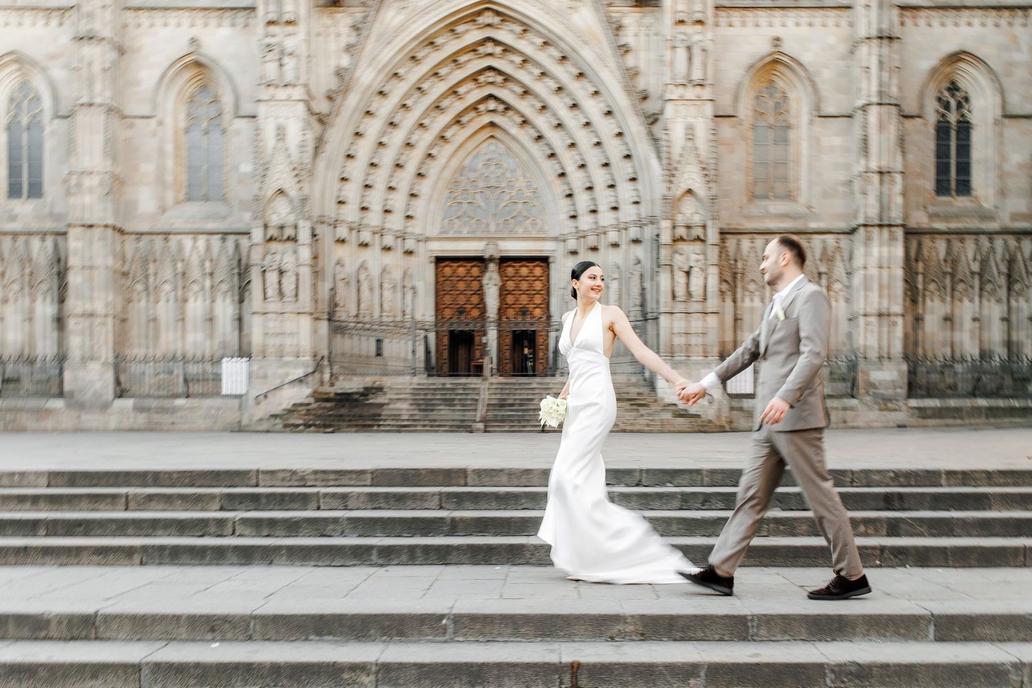 A couple in wedding attire walks in front of the Barcelona Cathedral. The bride holds a bouquet as they look at each other.