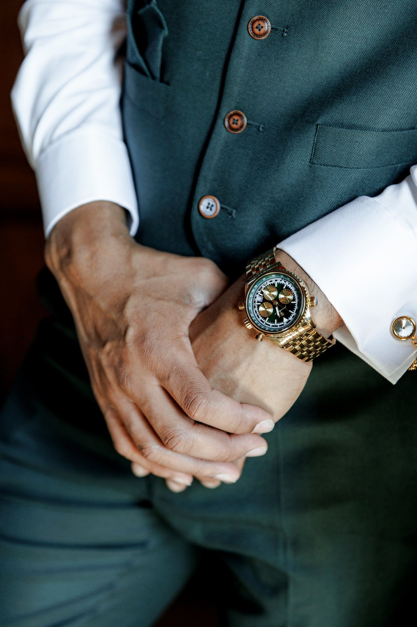 Groom adjusting gold skeleton watch with cufflinks, wearing white dress shirt and green suit vest.