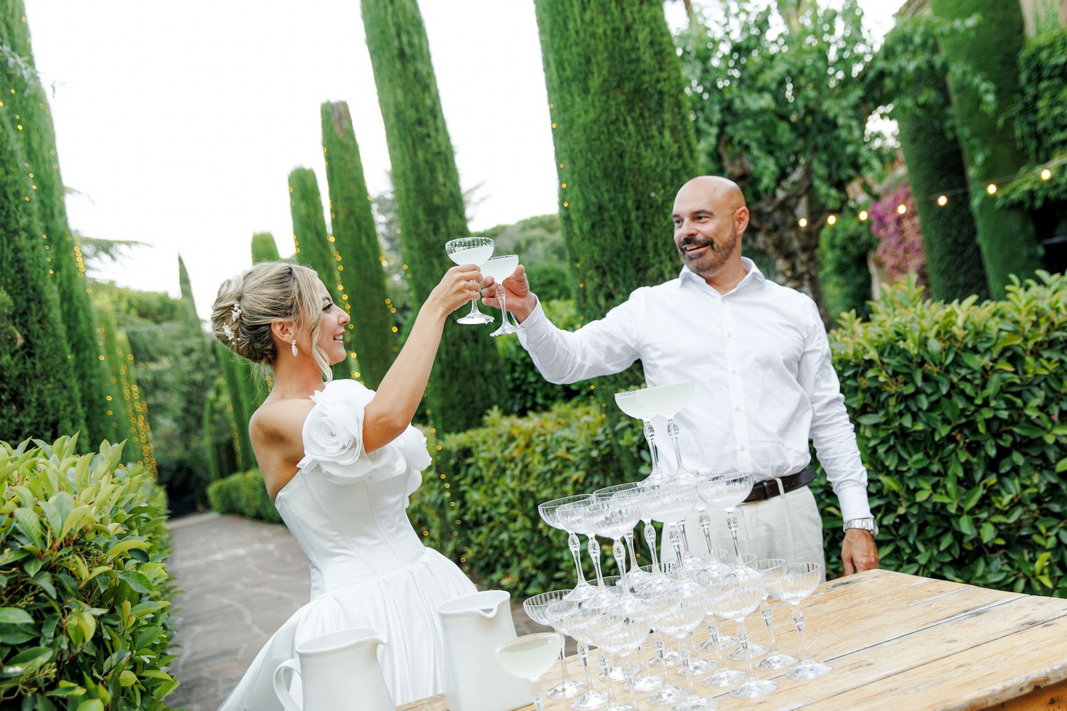 Bride and groom toasting champagne glasses outdoors at elegant garden wedding with cypress trees.