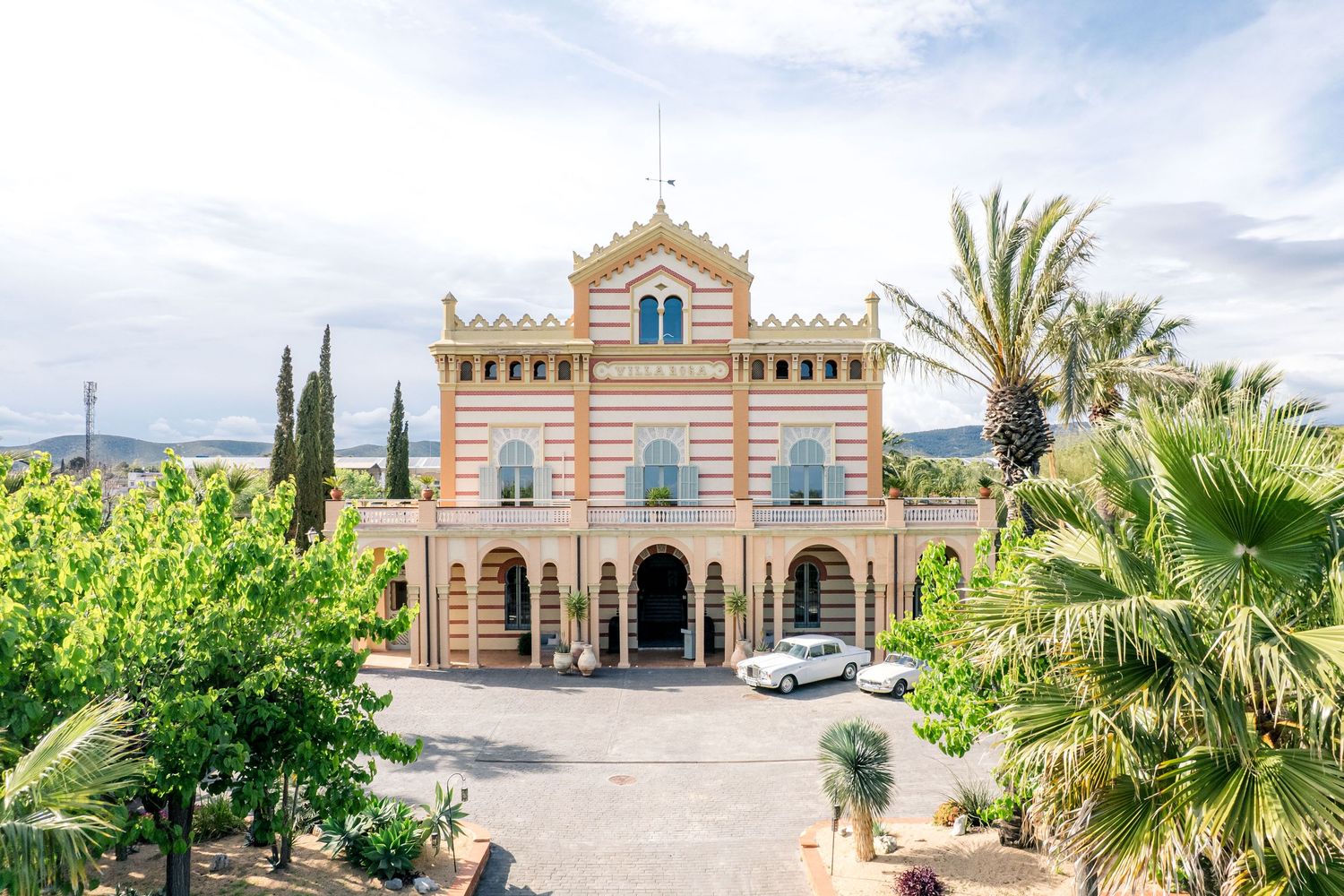 Elegant pink Mediterranean villa with palm trees, arched entrance, and manicured grounds under a cloudy sky.