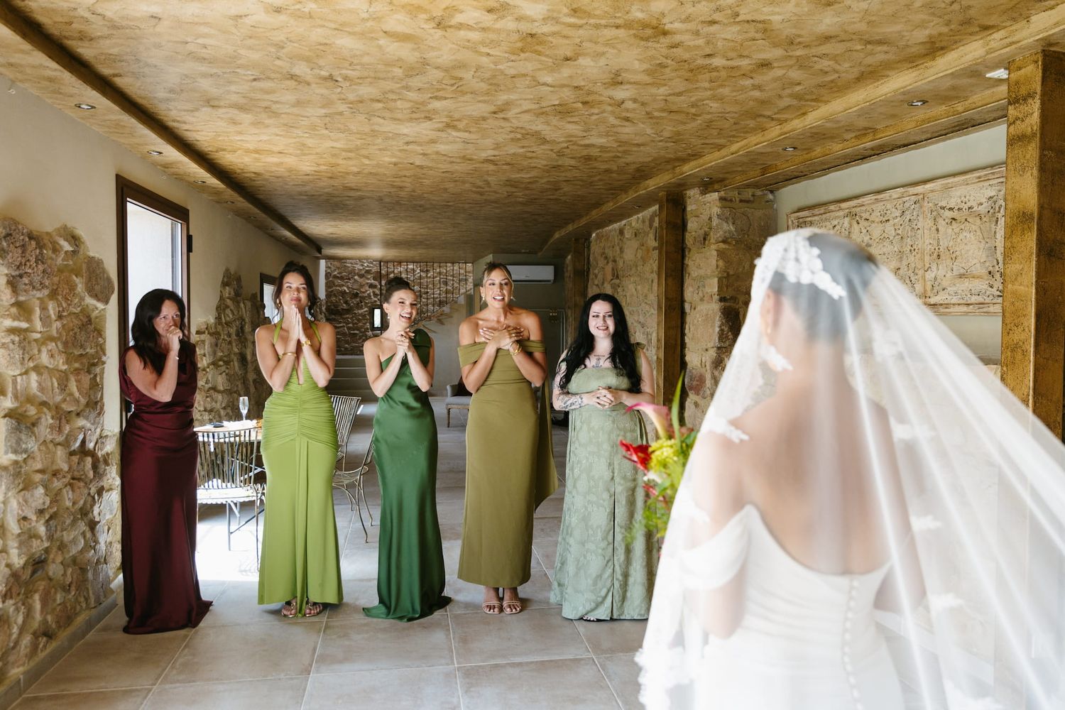 Bridesmaids in green and burgundy dresses react with joy as bride in white gown and veil reveals herself during first look.