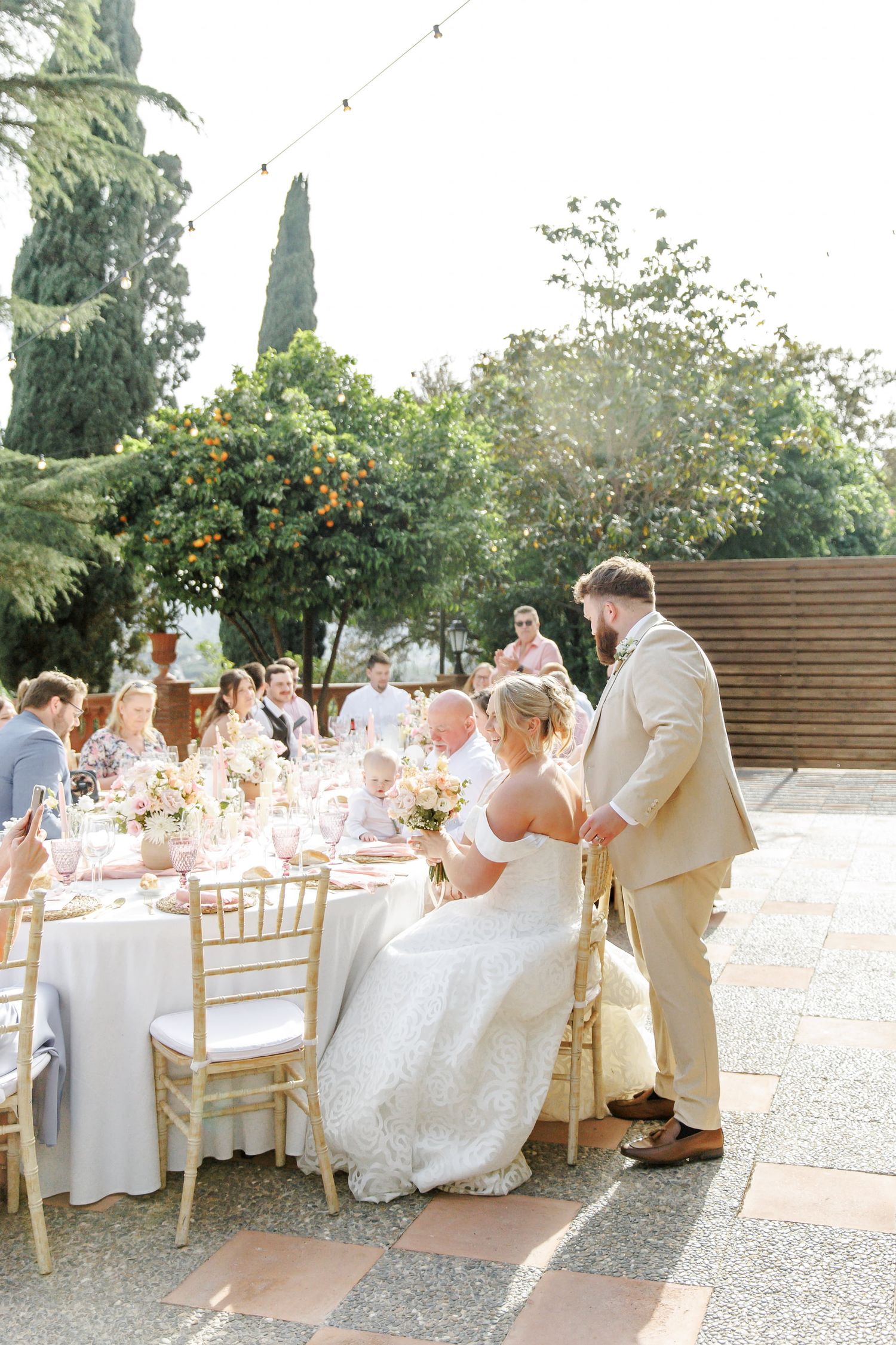 Bride and groom at elegant outdoor wedding reception with gold chairs, floral centerpieces, and lush greenery backdrop.