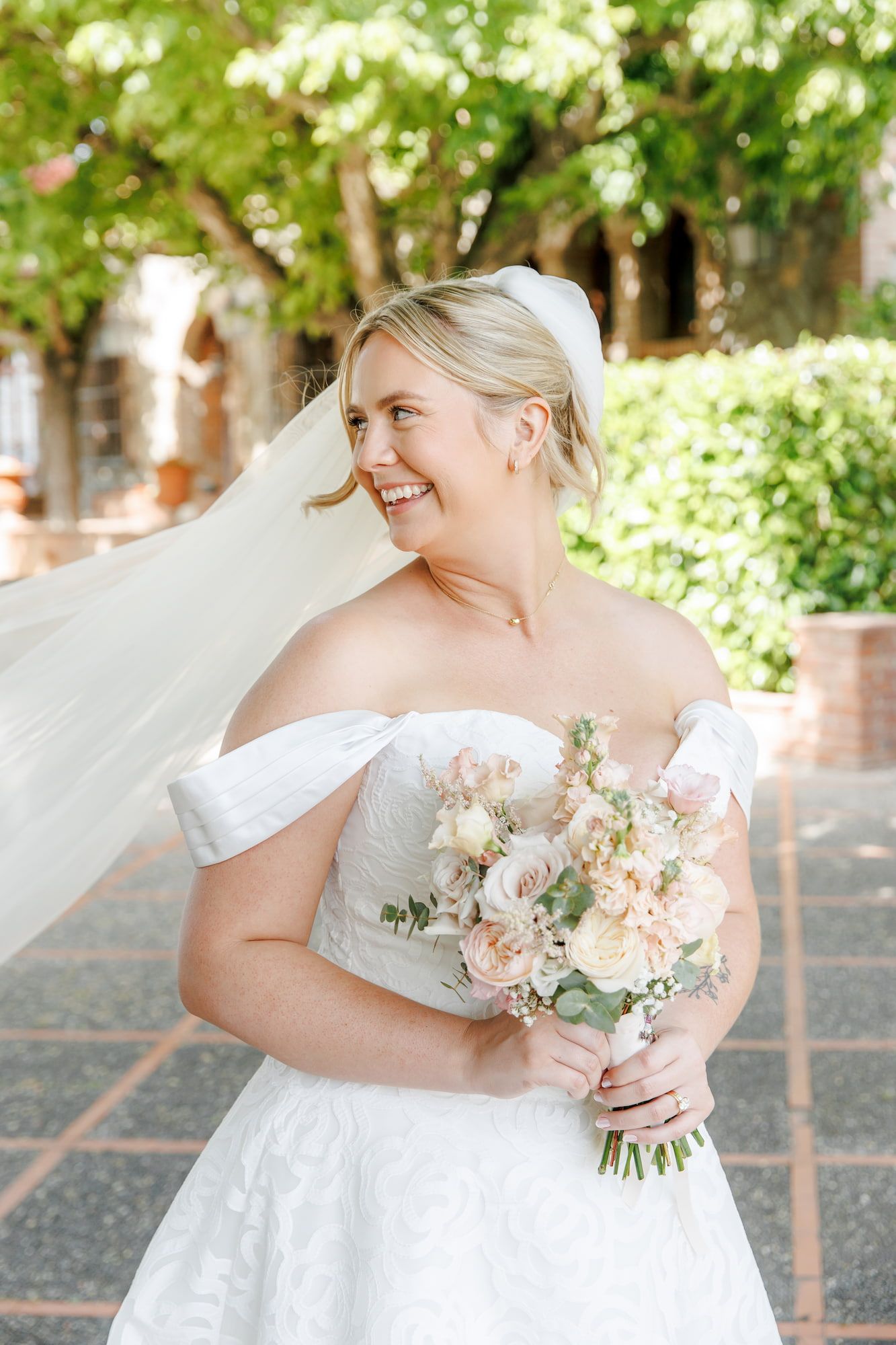 Smiling blonde bride in off-shoulder white gown holds blush bouquet, veil flowing in breeze outdoors.