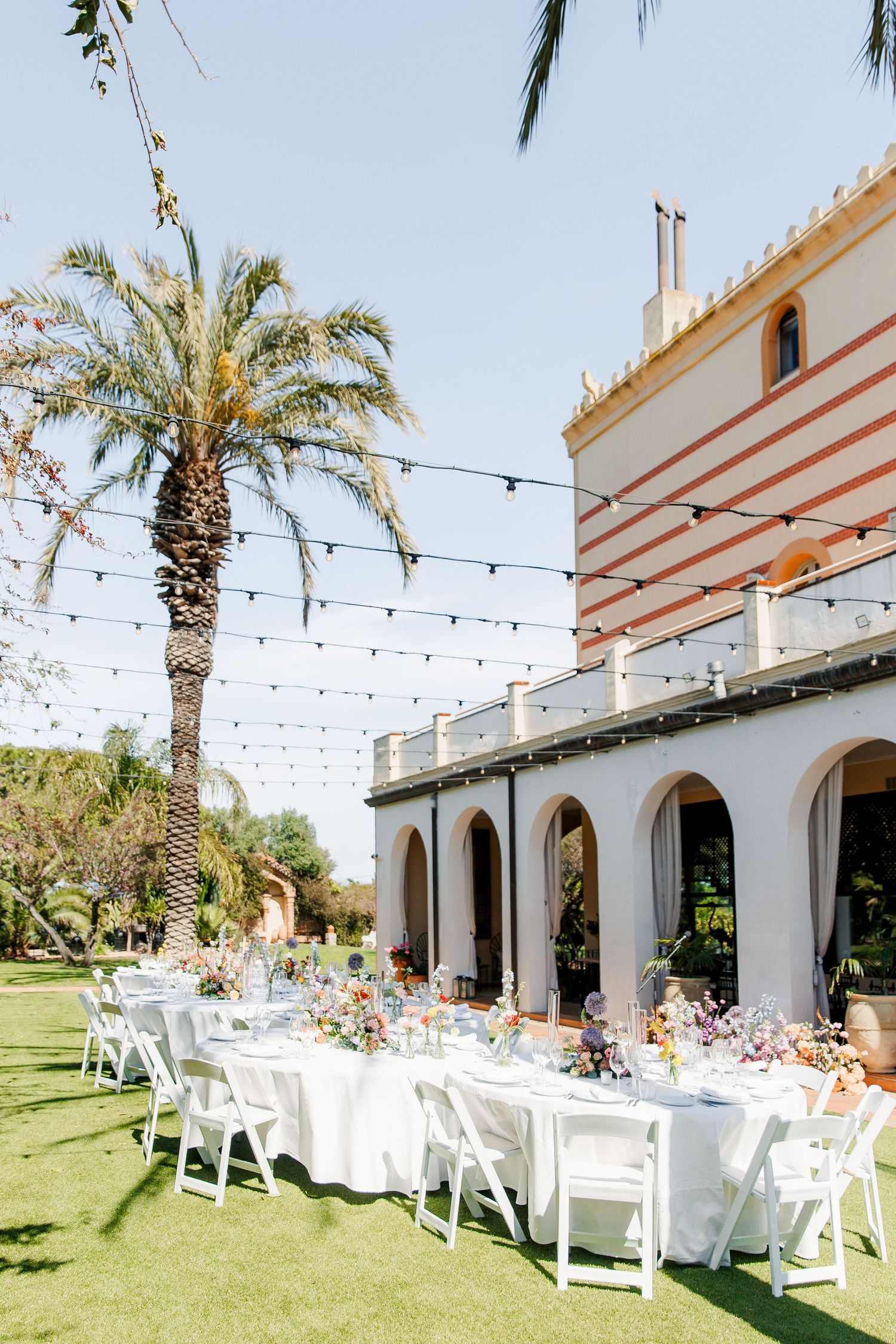 Outdoor wedding reception with white tables, chairs, string lights, palm trees, and a elegant striped building in sunny garden.