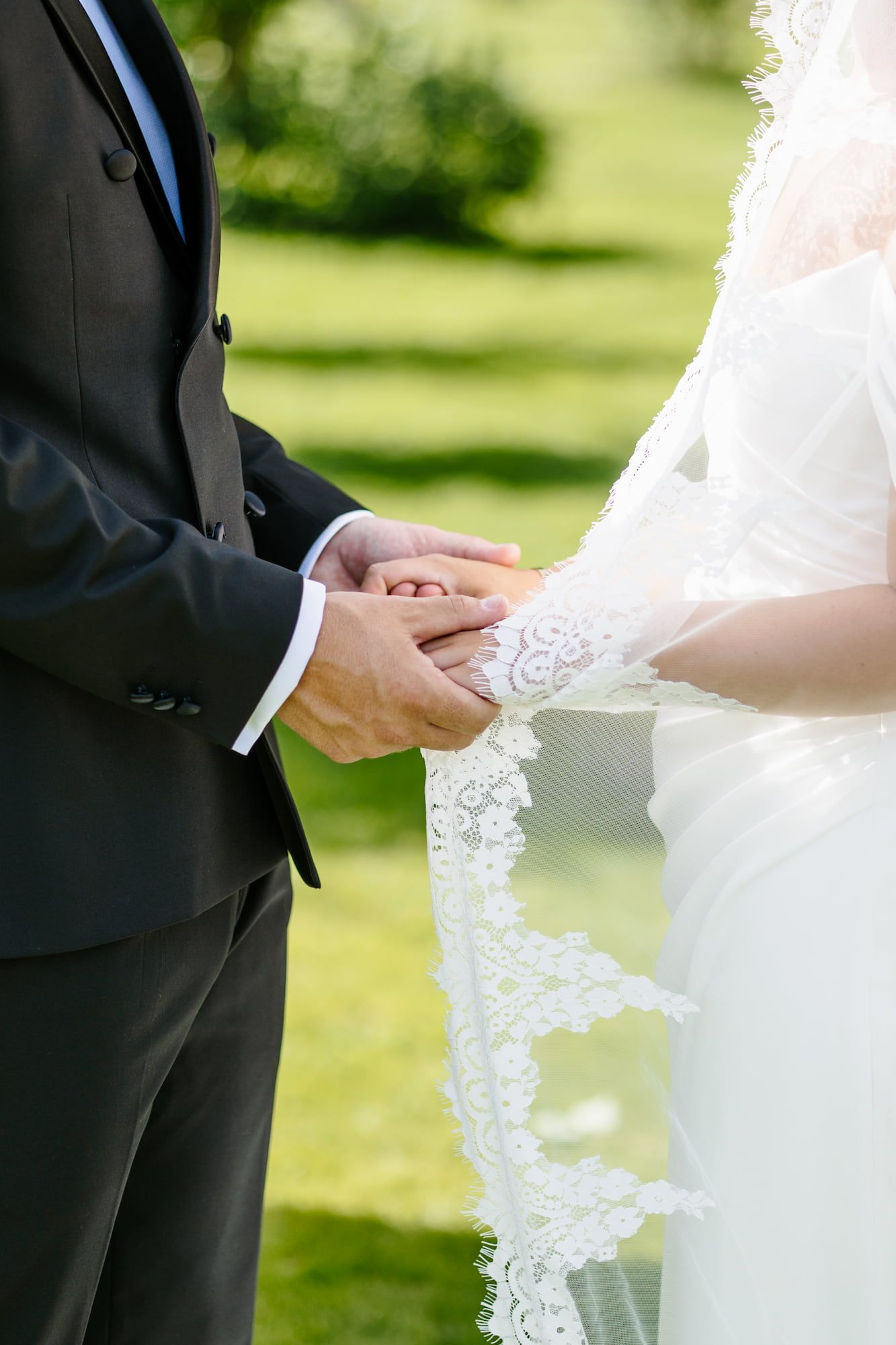 Bride and groom holding hands during outdoor wedding ceremony, bride wearing lace veil and gown, groom in dark suit.