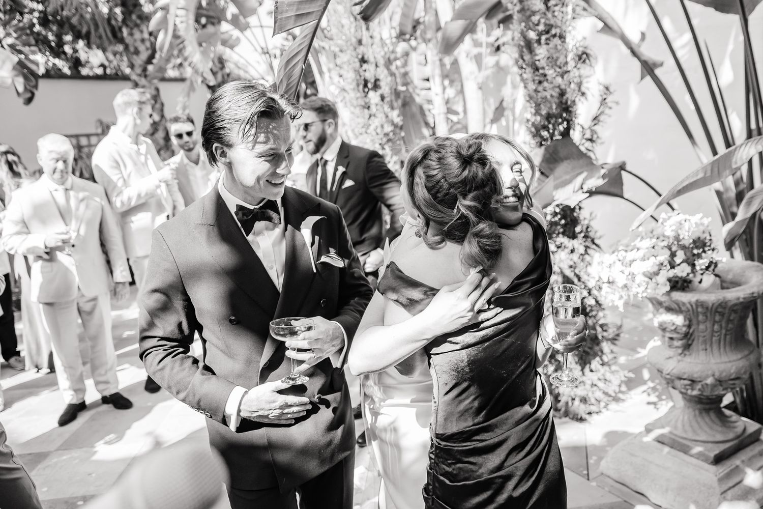 Black and white wedding photo of groom in tuxedo sharing a joyful moment with guests near a floral arrangement.