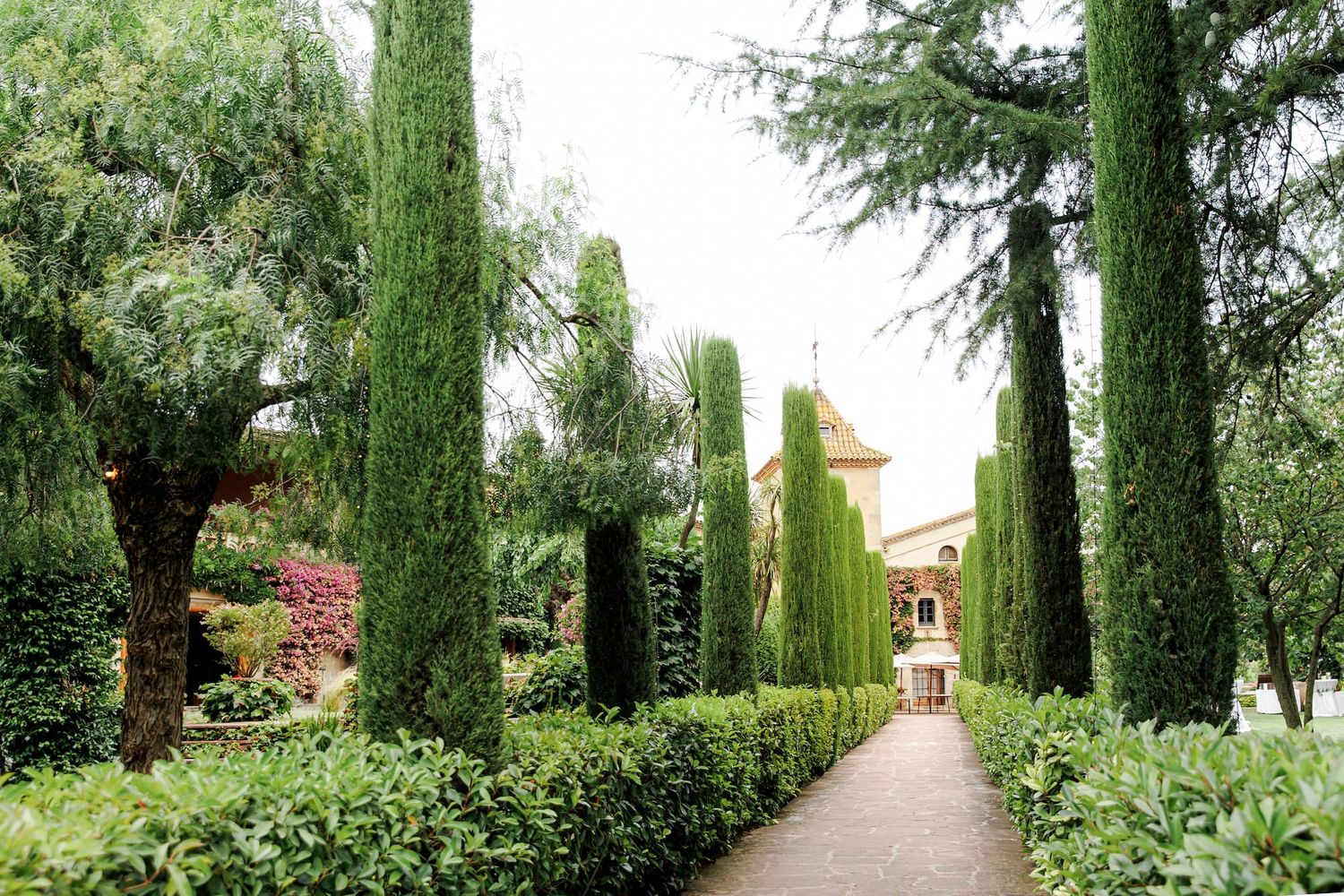 Green garden path lined with tall cypress trees and manicured hedges leading to a wedding venue near Barcelona.