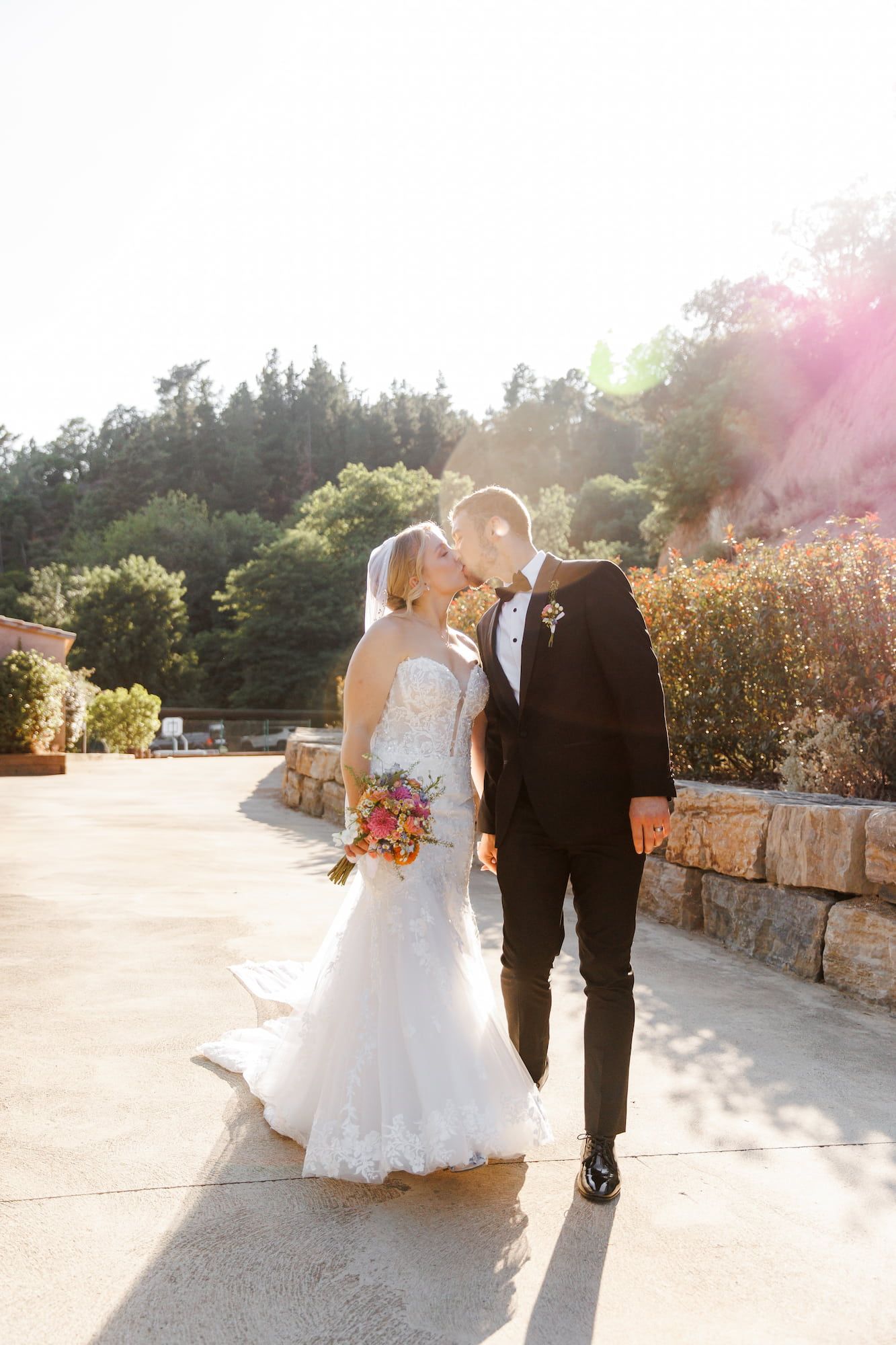 A romantic outdoor wedding scene with sunlit stone walls and lush greenery in the background.