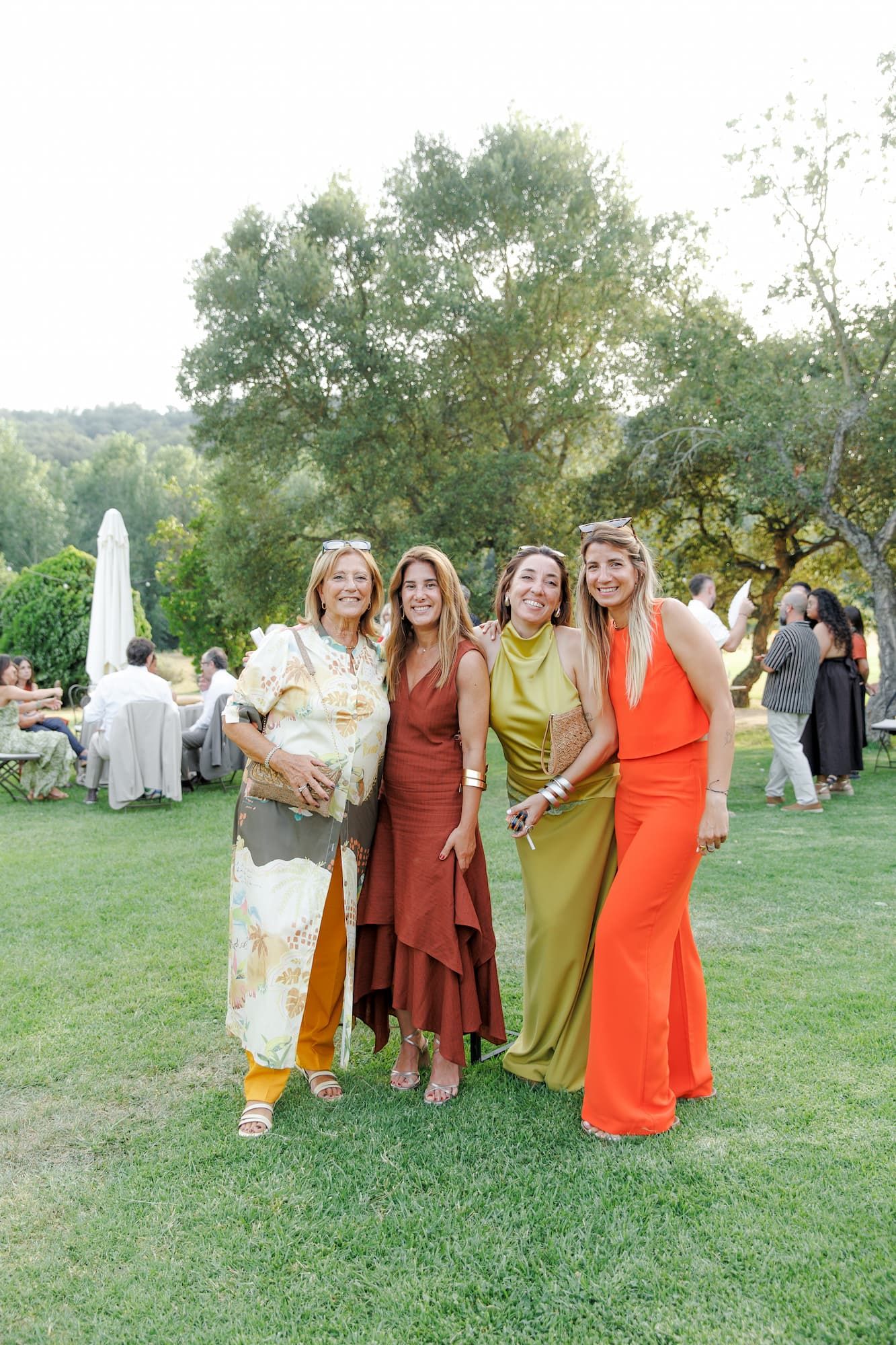 Four women in colorful formal dresses pose together on green grass at an outdoor garden party event.