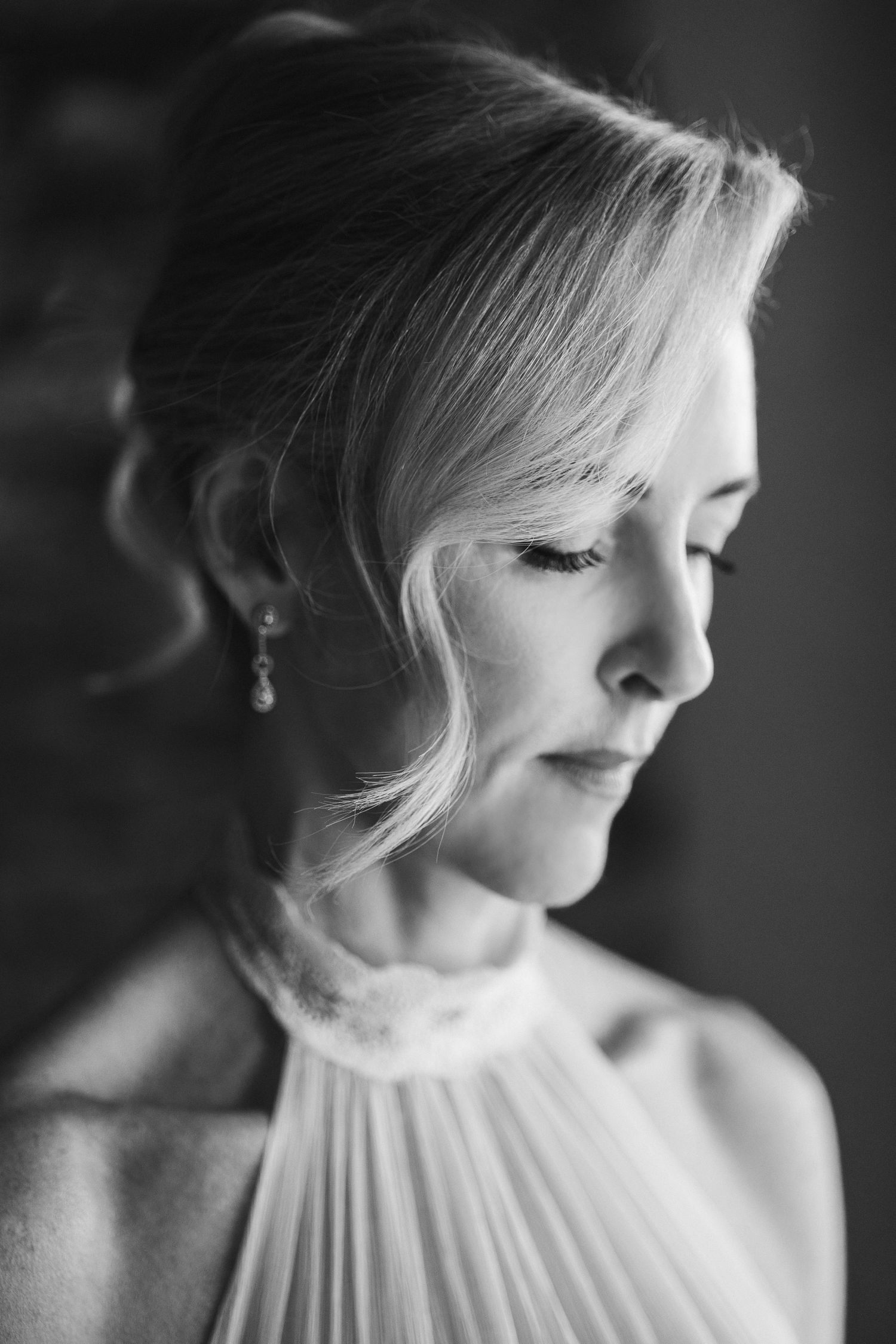 Black and white profile portrait of a woman with an elegant updo and drop earring, eyes gently closed, wearing a pleated halter-neck gown.