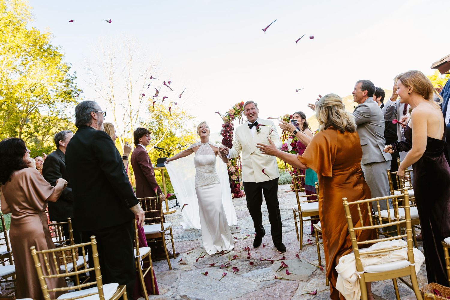 Bride and groom walk down the aisle outdoors as seated wedding guests cheer and toss petals in the air during a joyful ceremony.