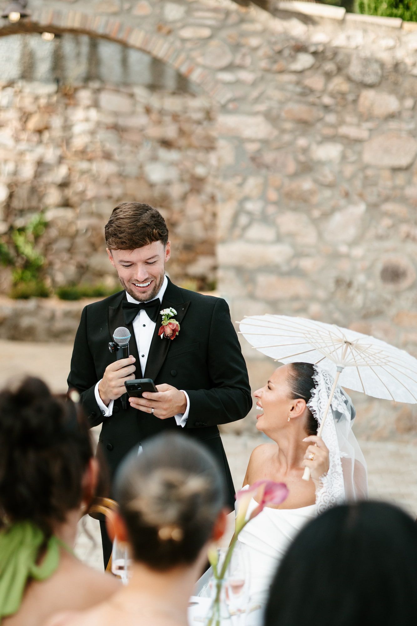 Groom in black tuxedo reads vows from phone while smiling bride holds white parasol at outdoor wedding ceremony.